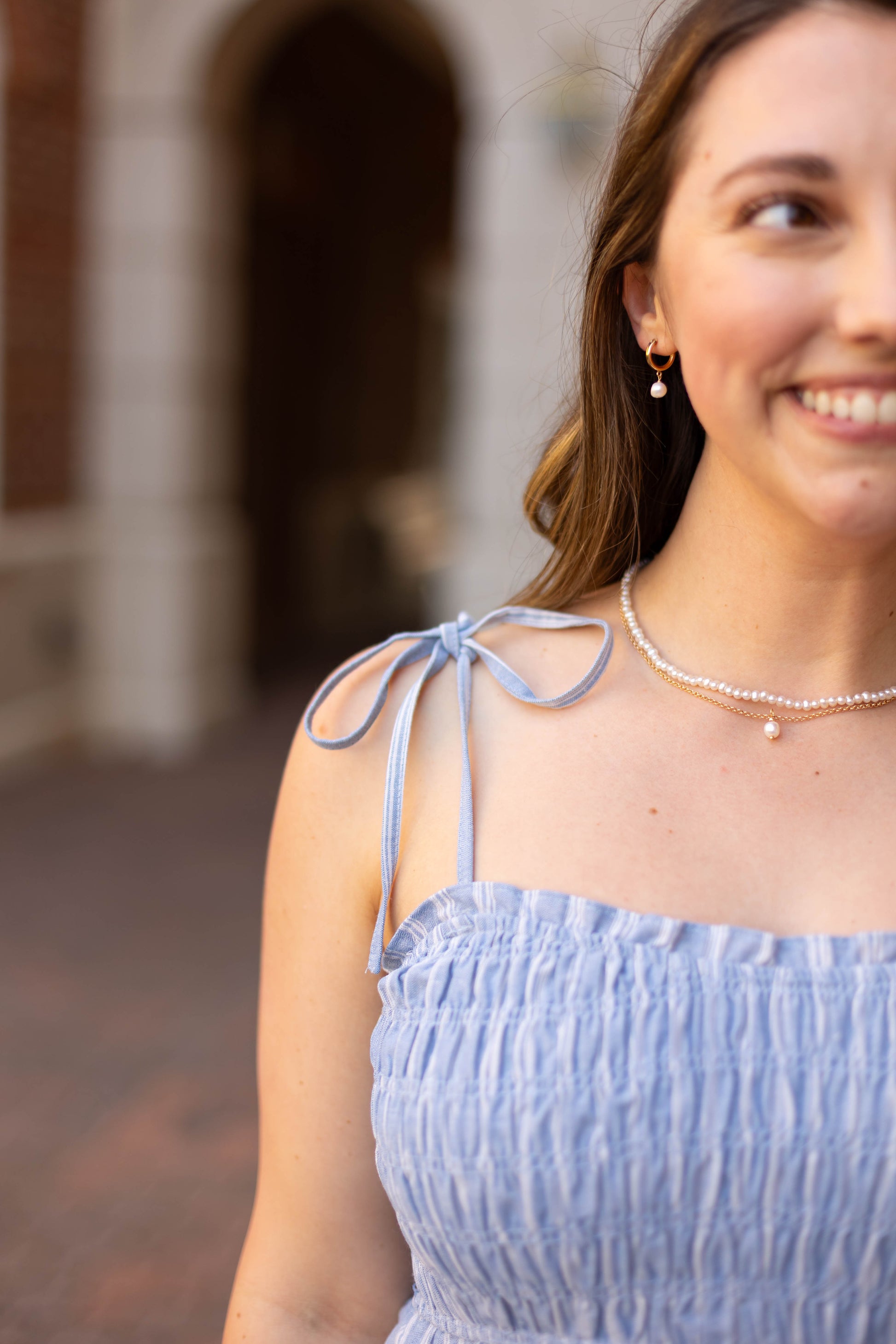 A woman smiles outdoors, wearing the Dogwood Cloth Amelia Striped Midi—a light blue, sleeveless, ruched 100% cotton dress with tie straps—paired with a pearl necklace and gold earrings; only the right side of her face is visible.