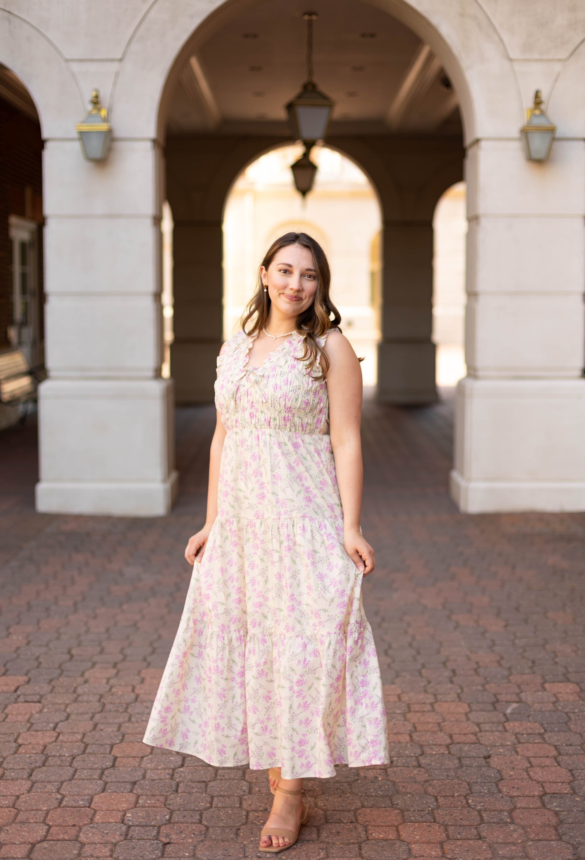 A woman wearing the Sophia Floral Midi by Dogwood Cloth, featuring a tiered silhouette, stands on a brick pathway under an archway and smiles at the camera.