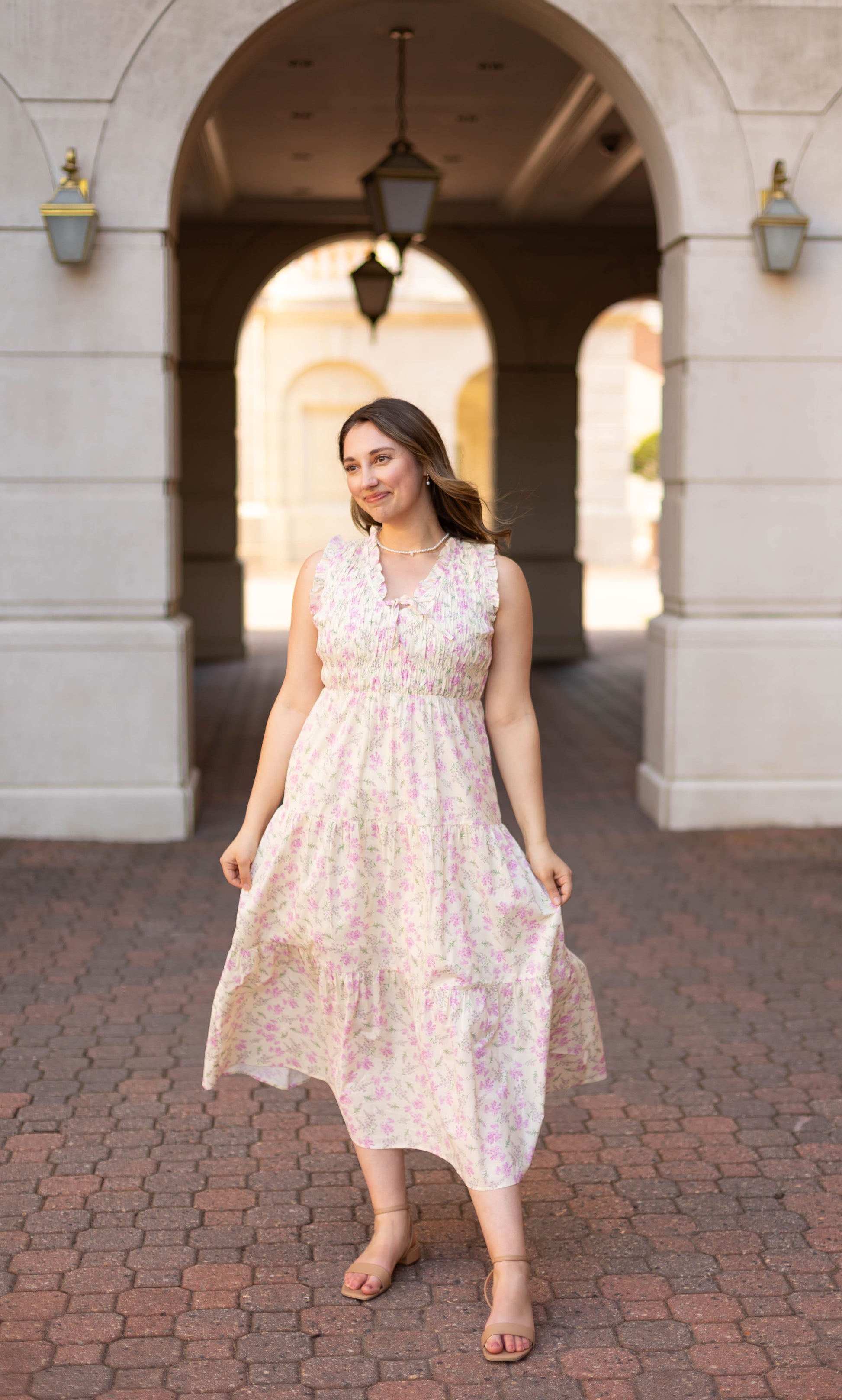 A woman in the Dogwood Cloth Sophia Floral Midi stands on a brick pathway under an archway, facing forward with a slight smile.