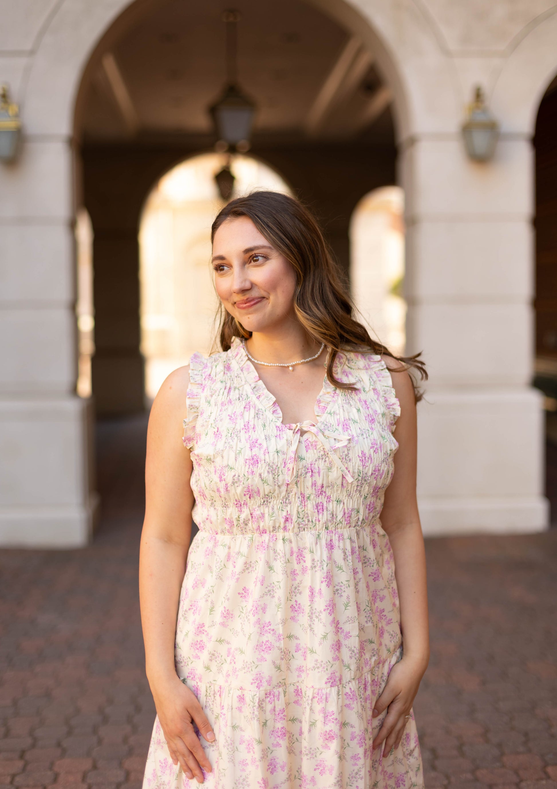 A woman wearing the Sophia Floral Midi by Dogwood Cloth stands outdoors before an arched stone building, gazing to the side with a smile.
