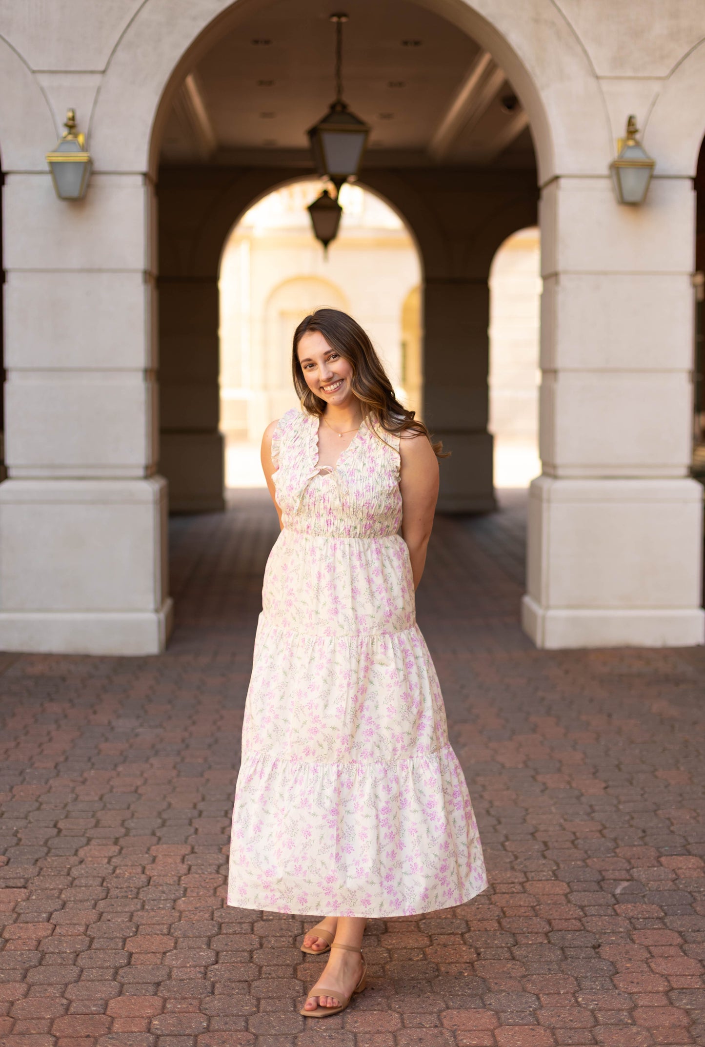 A woman smiles with her hands behind her back, standing on a brick walkway beneath an arched entrance, wearing the Sophia Floral Midi by Dogwood Cloth.