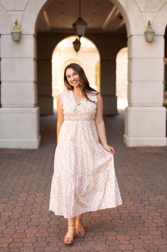 A woman in the Sophia Floral Midi by Dogwood Cloth stands on a brick walkway under an archway, holding one side of her dress and smiling at the camera.