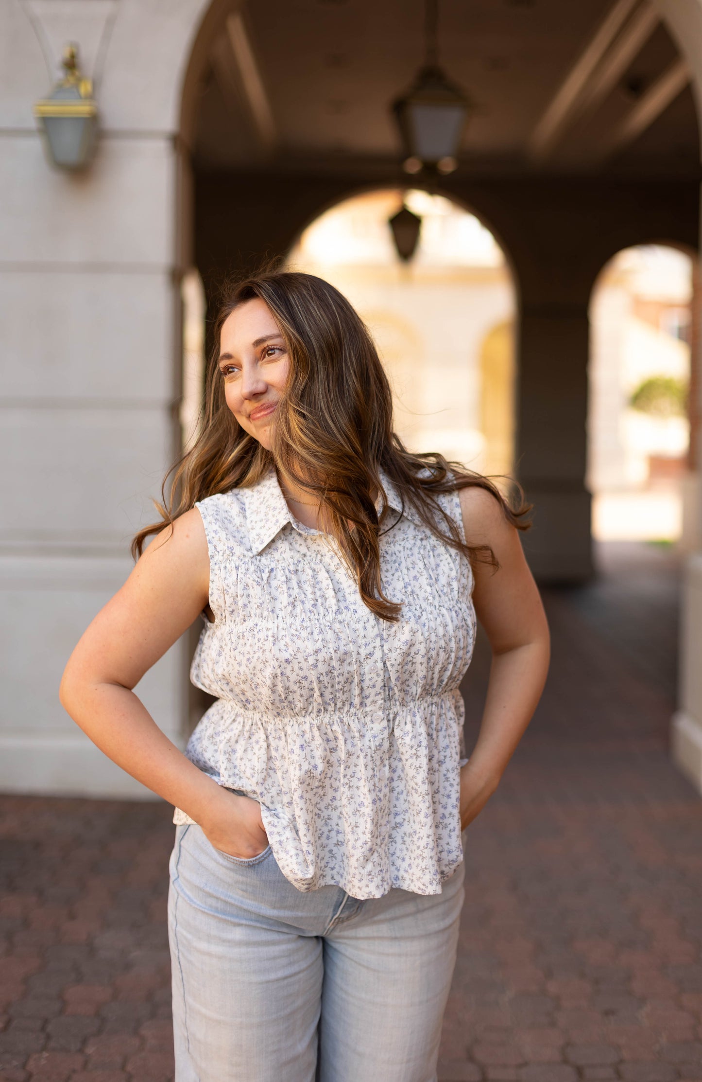 A woman with long brown hair, wearing the Dogwood Cloth Lily Floral Top and light jeans, stands outdoors with her hands in her pockets, smiling and looking to the side.