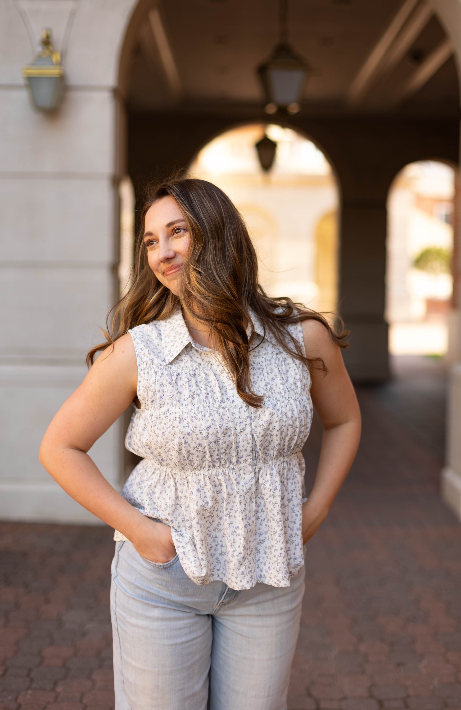 A woman with long brown hair, wearing the Dogwood Cloth Lily Floral Top and light jeans, stands outdoors with her hands in her pockets, smiling and looking to the side.