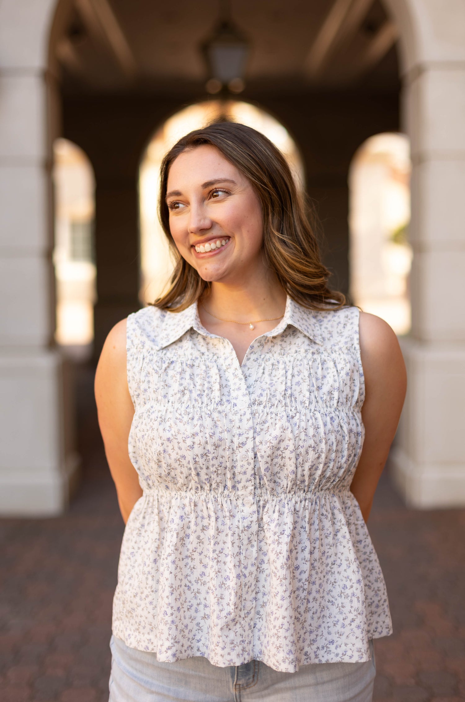 A woman with long brown hair smiles outdoors, wearing the Lily Floral Top by Dogwood Cloth—a sleeveless blouse with a subtle pattern. Elegant arched architecture forms the background.