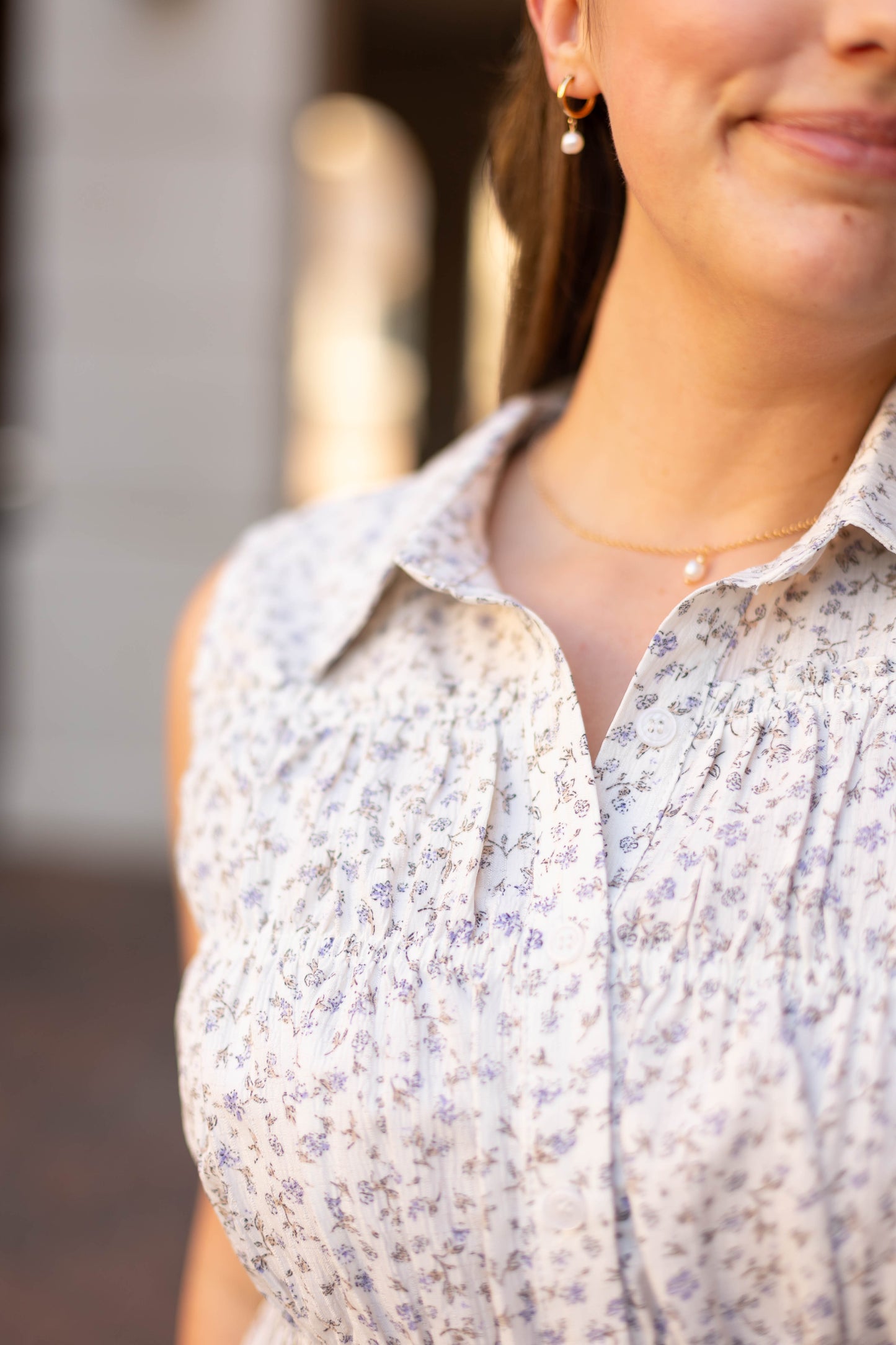 A woman, photographed from the shoulders up, wears the Dogwood Cloth Lily Floral Top—a light-colored, sleeveless blouse with delicate floral patterns—paired with a gold necklace featuring a small pendant.