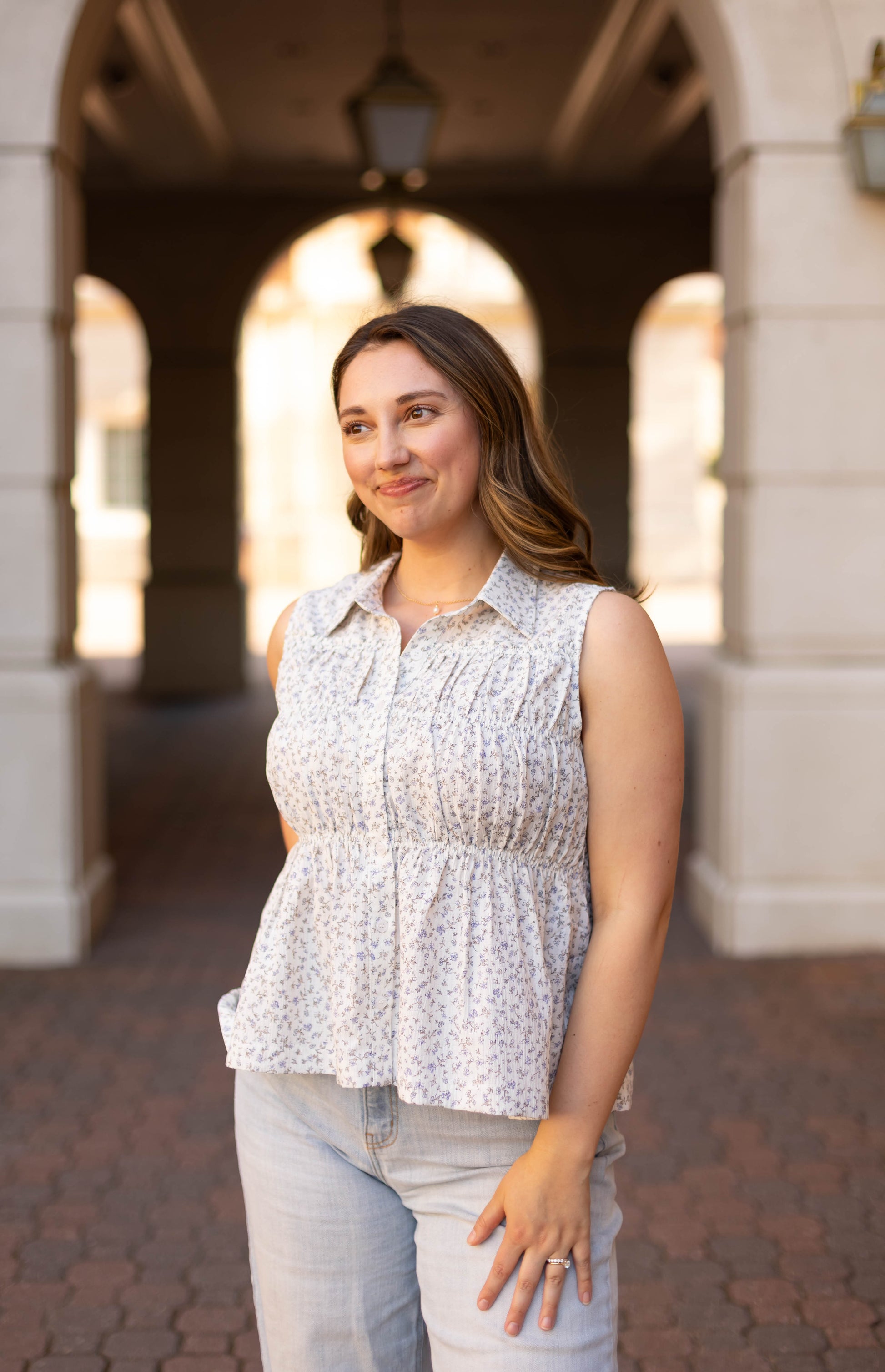 A woman with long hair smiles outdoors under an archway, wearing the Dogwood Cloth Lily Floral Top and light jeans, looking slightly to the side.