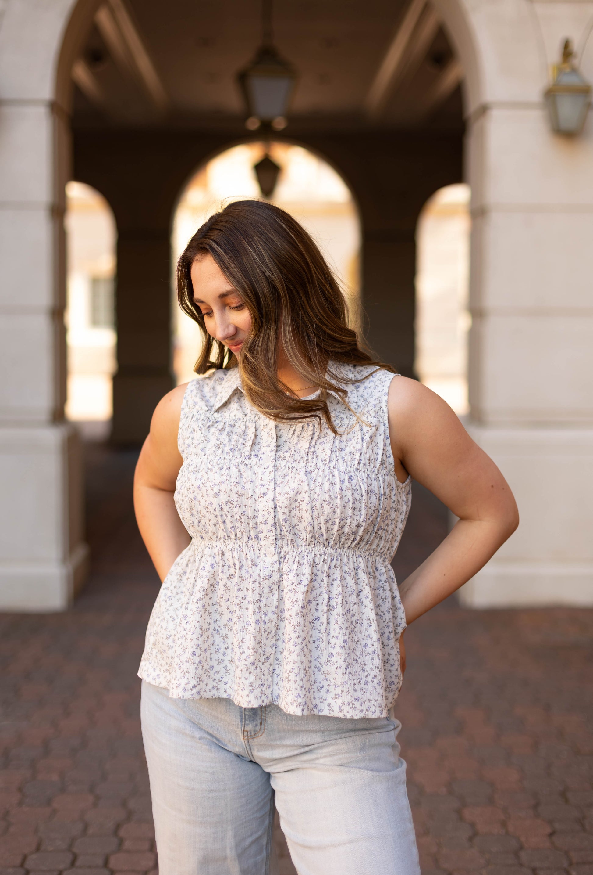 A woman with long brown hair stands outdoors, looking down with her hands on her lower back, wearing jeans and the Dogwood Cloth Lily Floral Top.