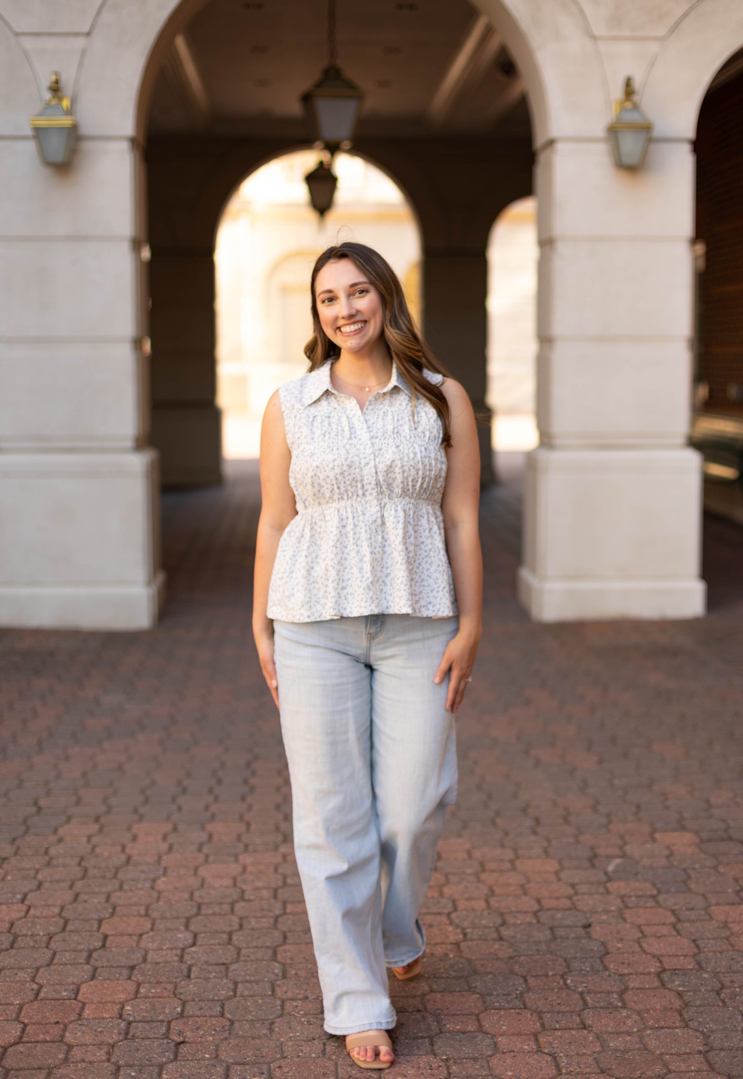 A woman in a sleeveless Lily Floral Top by Dogwood Cloth and light jeans smiles under an archway on a brick walkway, her outfit adding charm to the scene.
