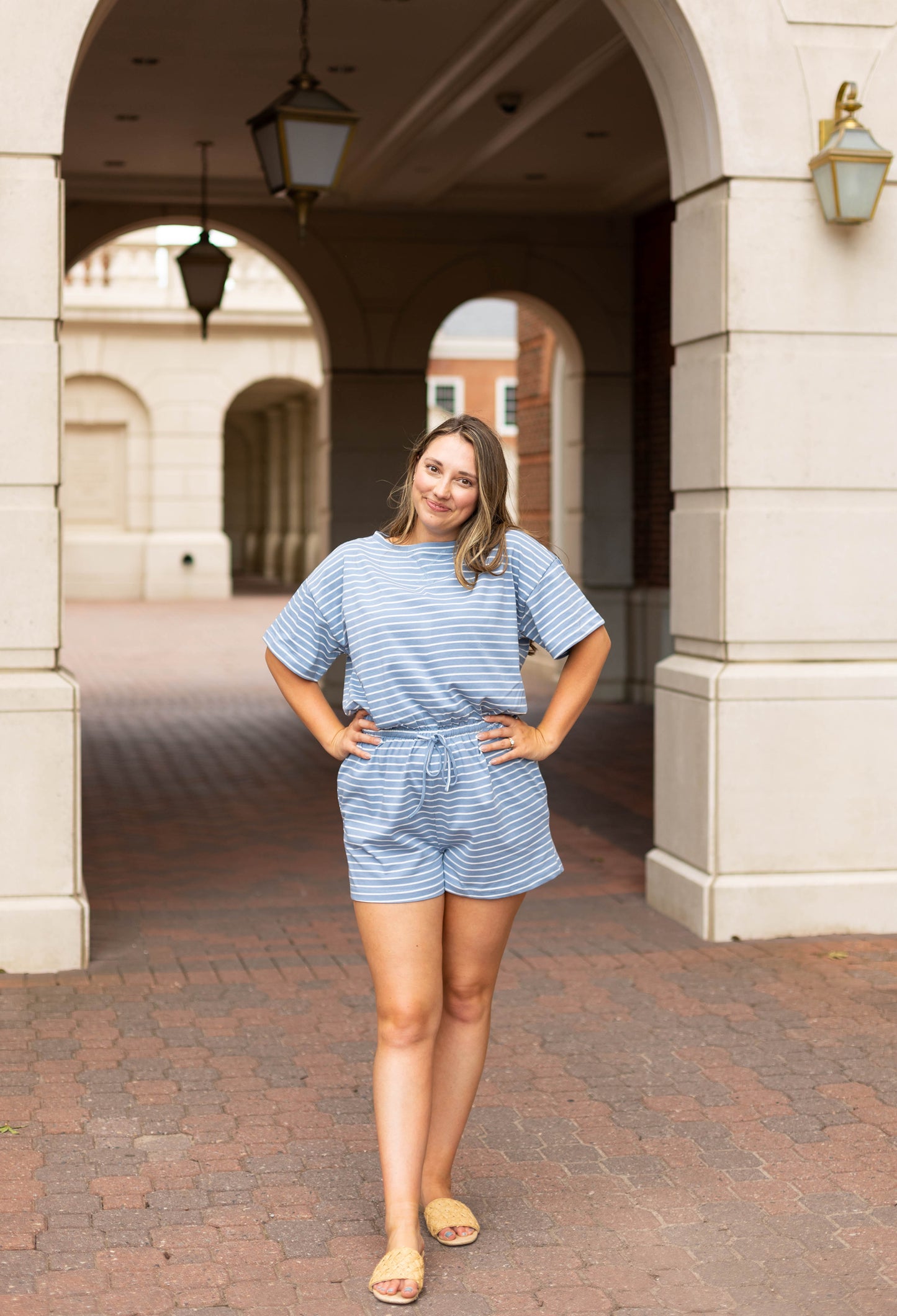 A woman stands with hands on hips under a stone archway, wearing the Dogwood Cloth Lucia Striped Romper and sandals. The blue and white stripes of the romper stand out against the brick pavement and buildings behind her.