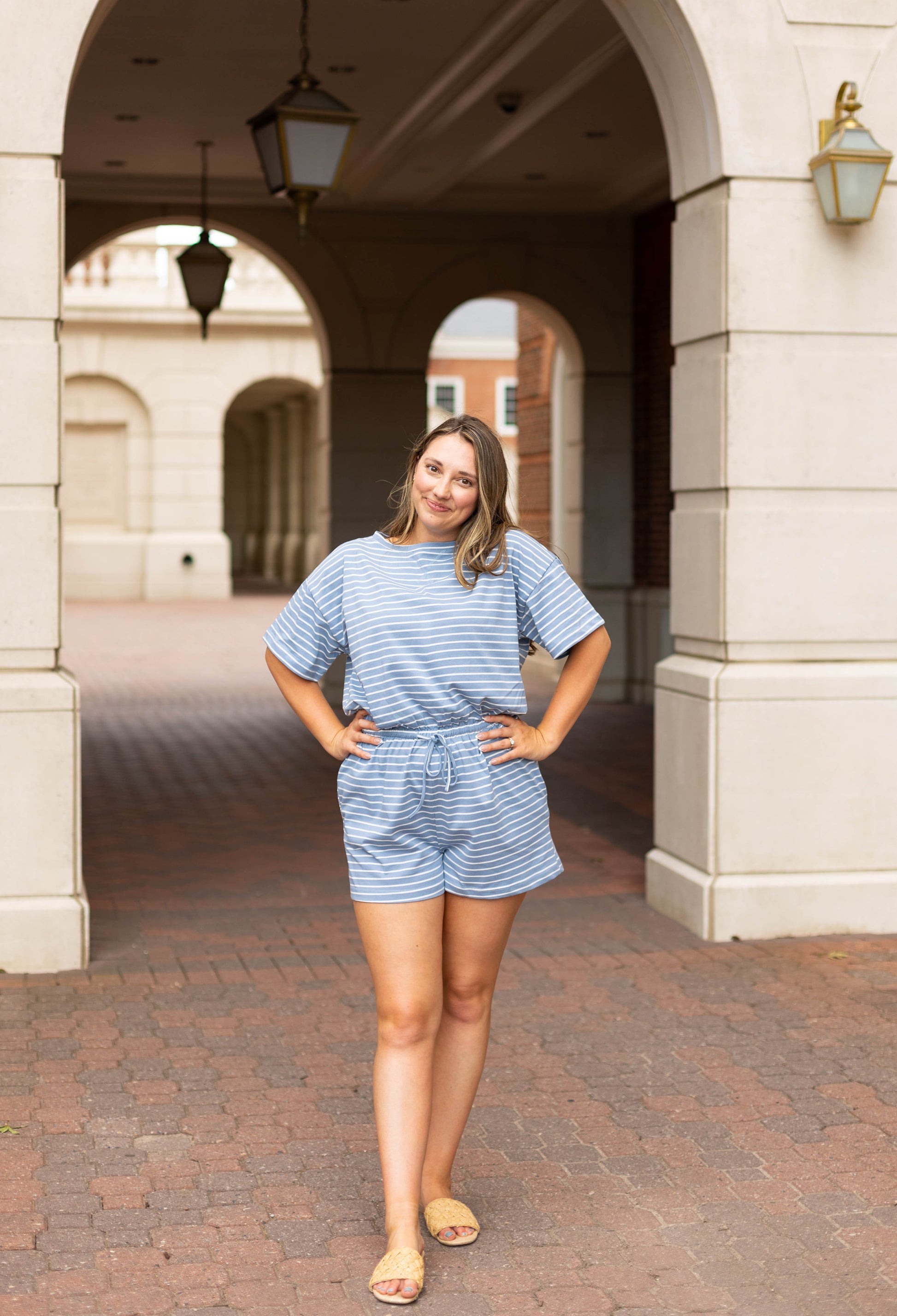 A woman stands with hands on hips under a stone archway, wearing the Dogwood Cloth Lucia Striped Romper and sandals. The blue and white stripes of the romper stand out against the brick pavement and buildings behind her.