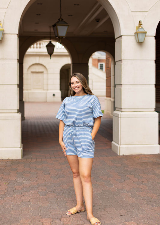 Young woman in the Dogwood Cloth Lucia Striped Romper smiles confidently under a stone archway with lanterns, standing on a brick walkway and showcasing this stylish women’s romper.
