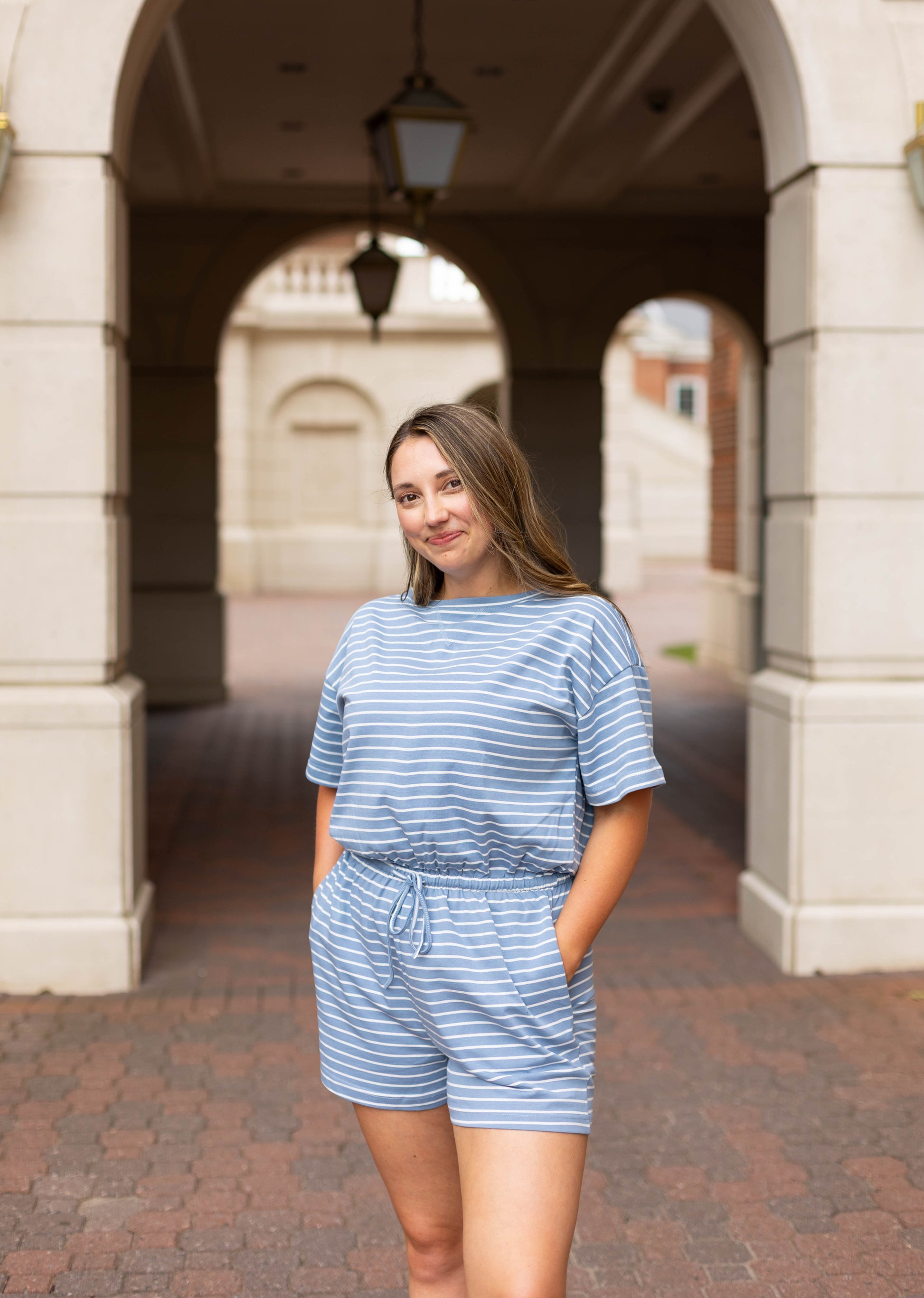 A woman wearing the Dogwood Cloth Lucia Striped Romper stands outdoors with her hands in her pockets, posing under a stone archway.