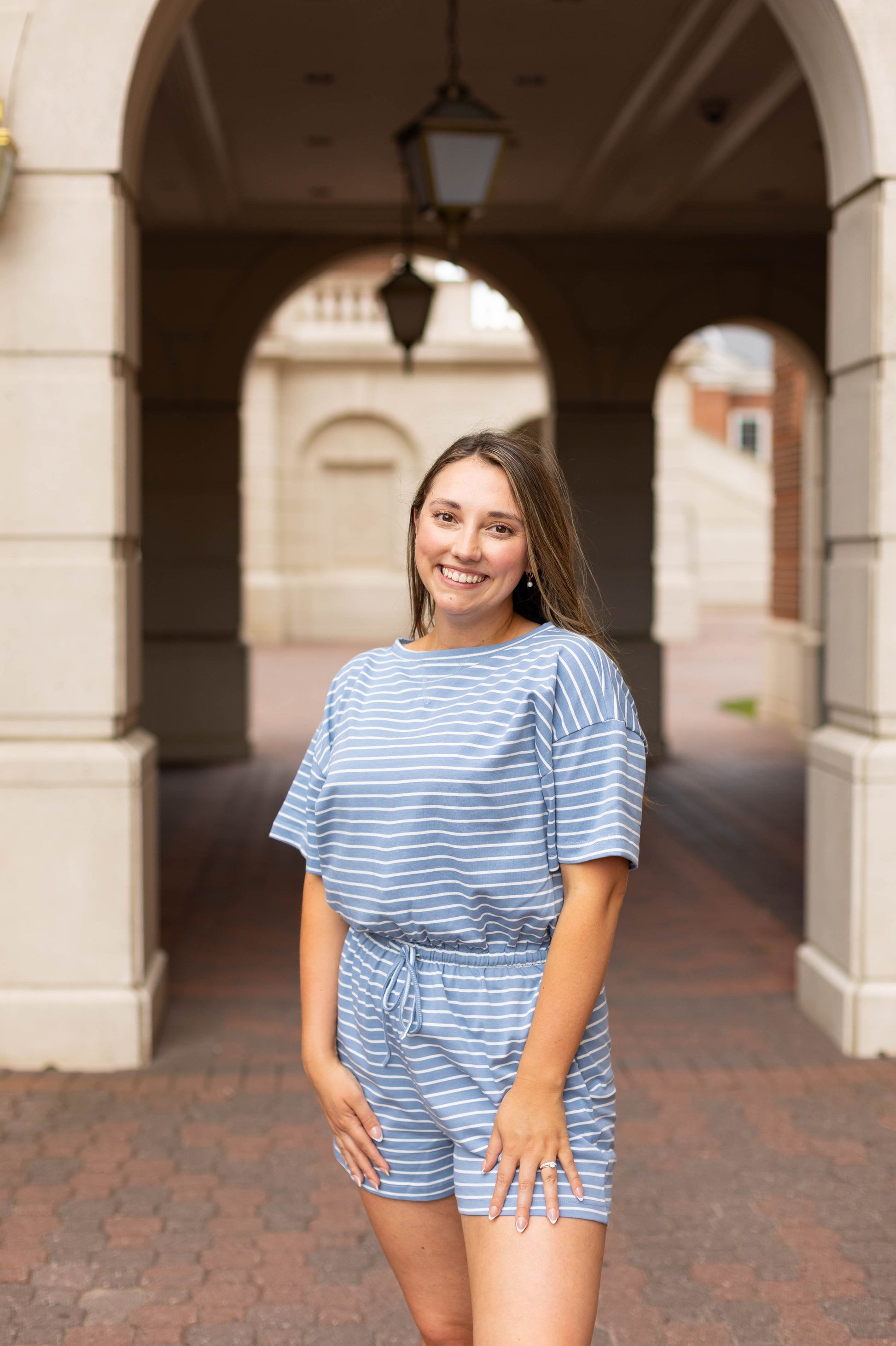 A young woman wears a blue and white Lucia Striped Romper by Dogwood Cloth, smiling in front of a building with arches and hanging lanterns.