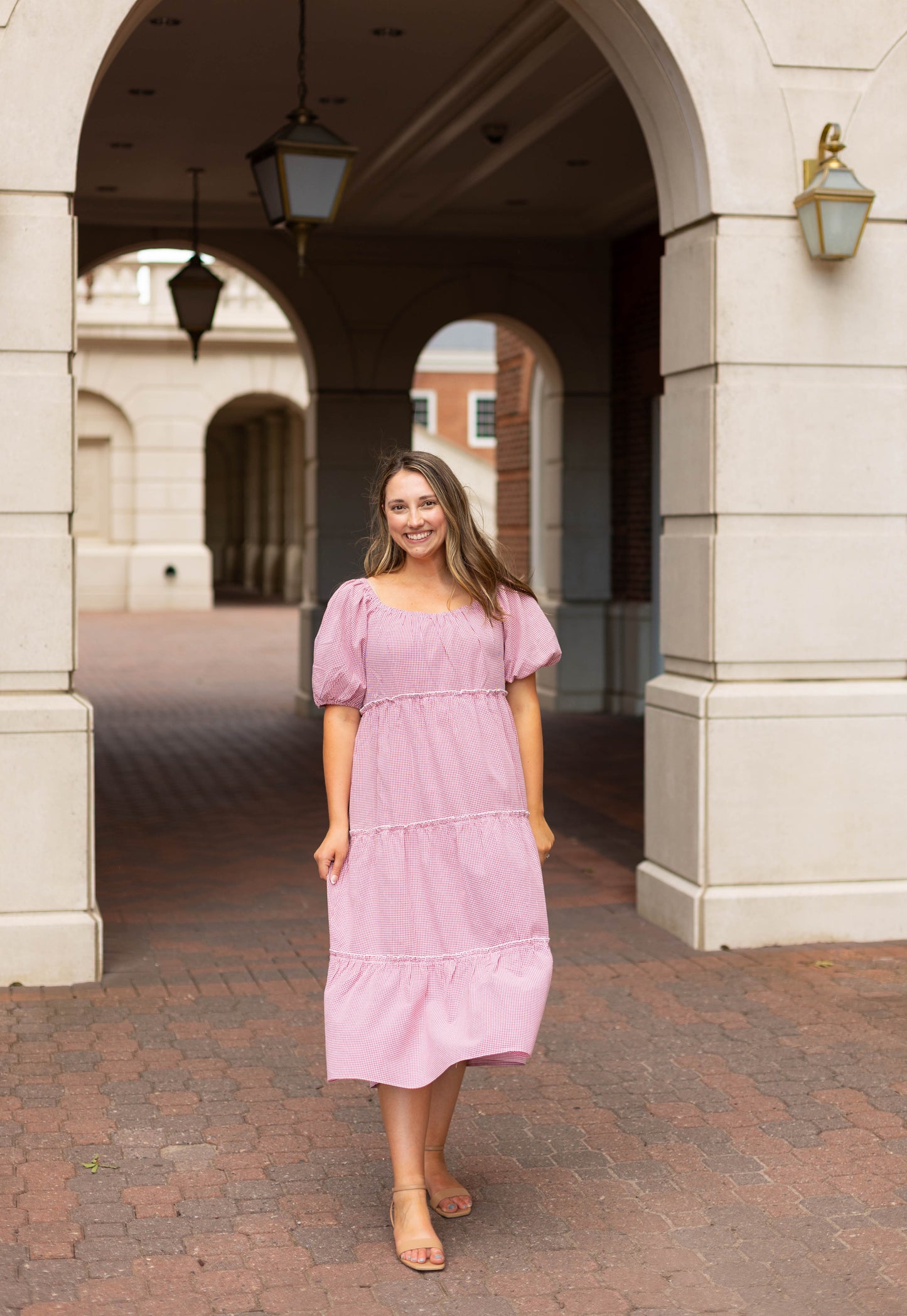 A woman smiles at the camera, standing on a brick walkway under a stone archway, wearing the Dogwood Cloth Courtlyn Tiered Gingham Midi.
