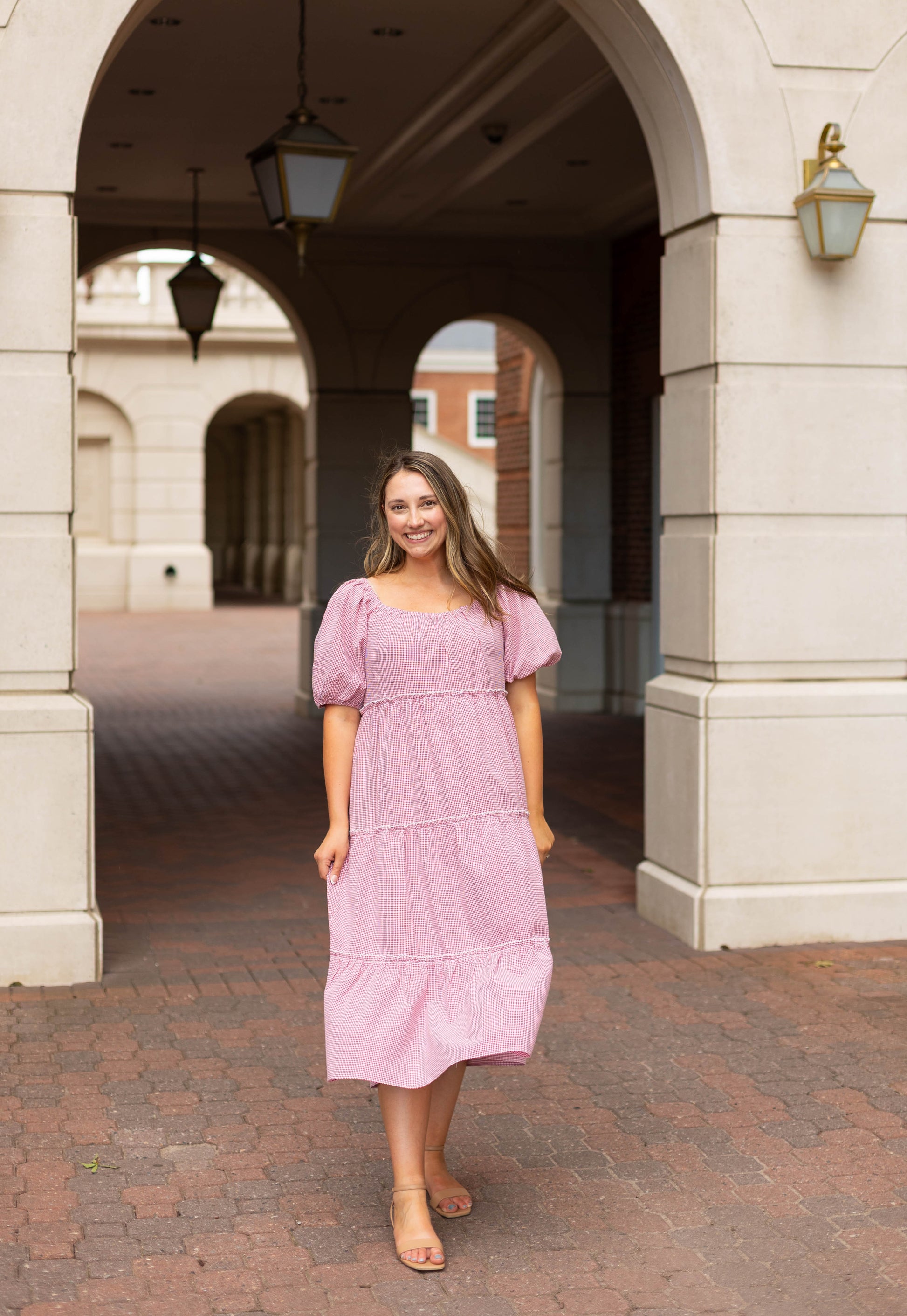 A woman smiles at the camera, standing on a brick walkway under a stone archway, wearing the Dogwood Cloth Courtlyn Tiered Gingham Midi.
