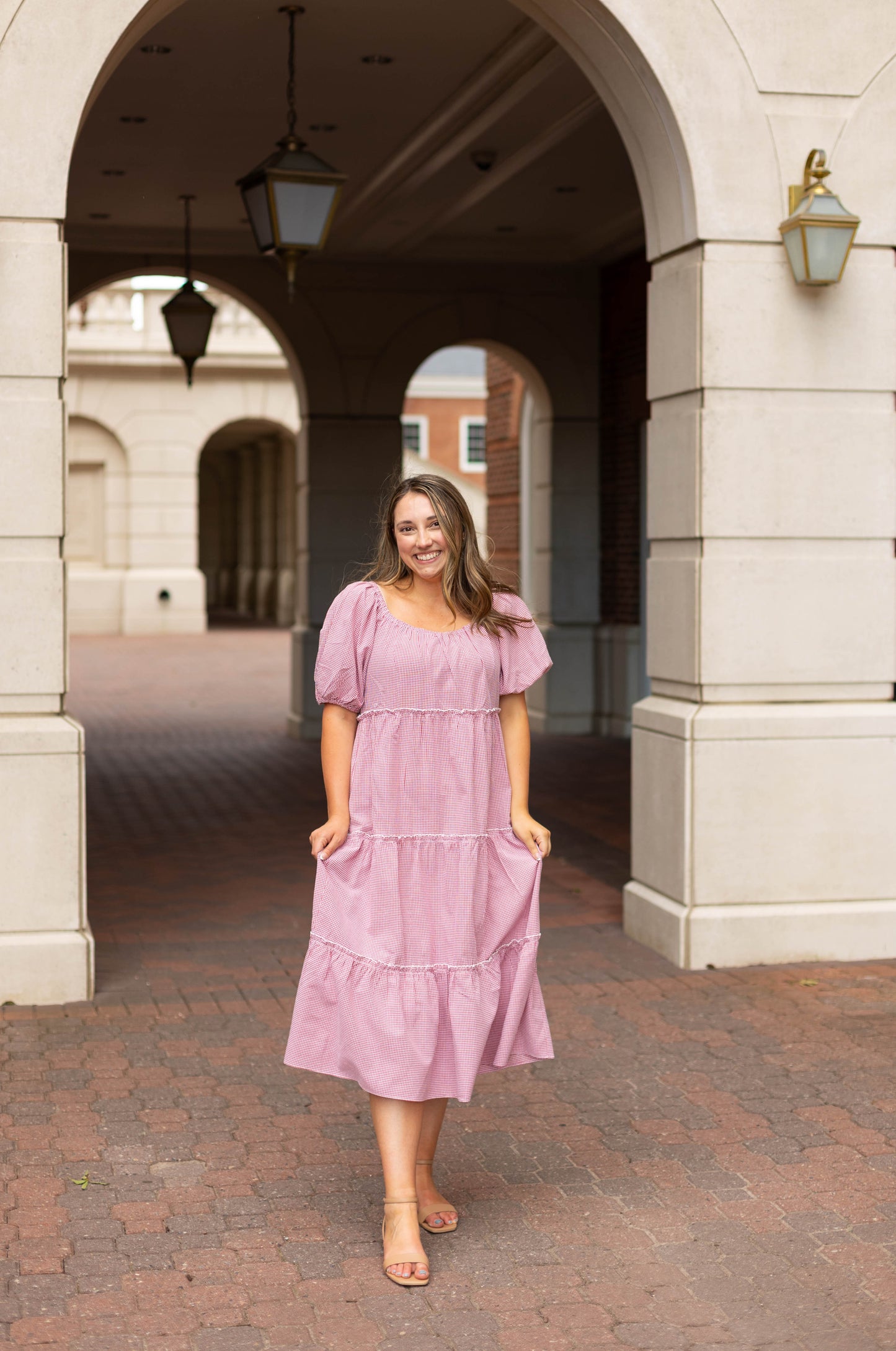 A woman stands outdoors under an archway, smiling at the camera while wearing the Dogwood Cloth Courtlyn Tiered Gingham Midi dress and sandals.