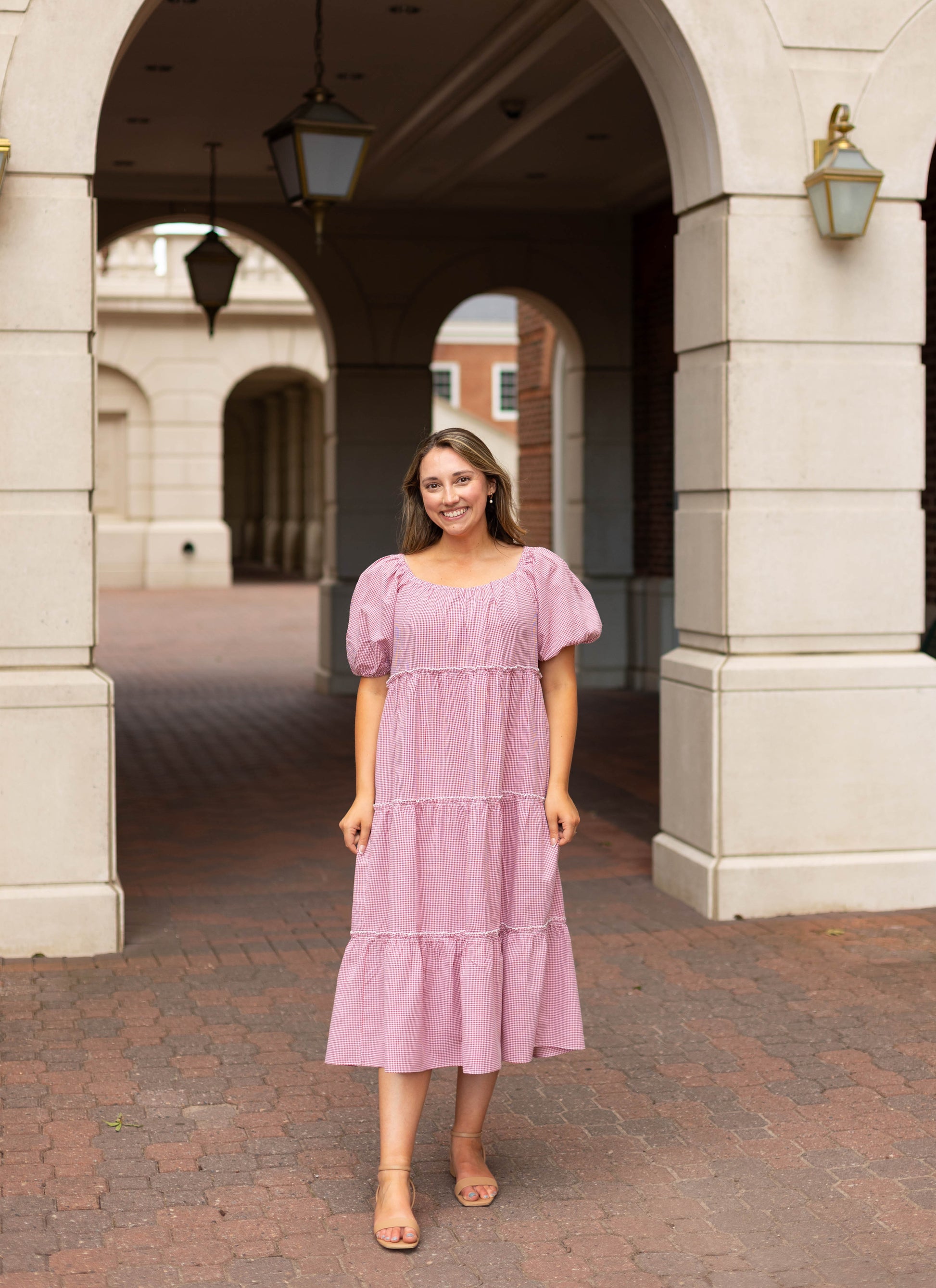 A woman in the Courtlyn Tiered Gingham Midi by Dogwood Cloth and tan sandals smiles under an archway on a brick walkway outdoors.