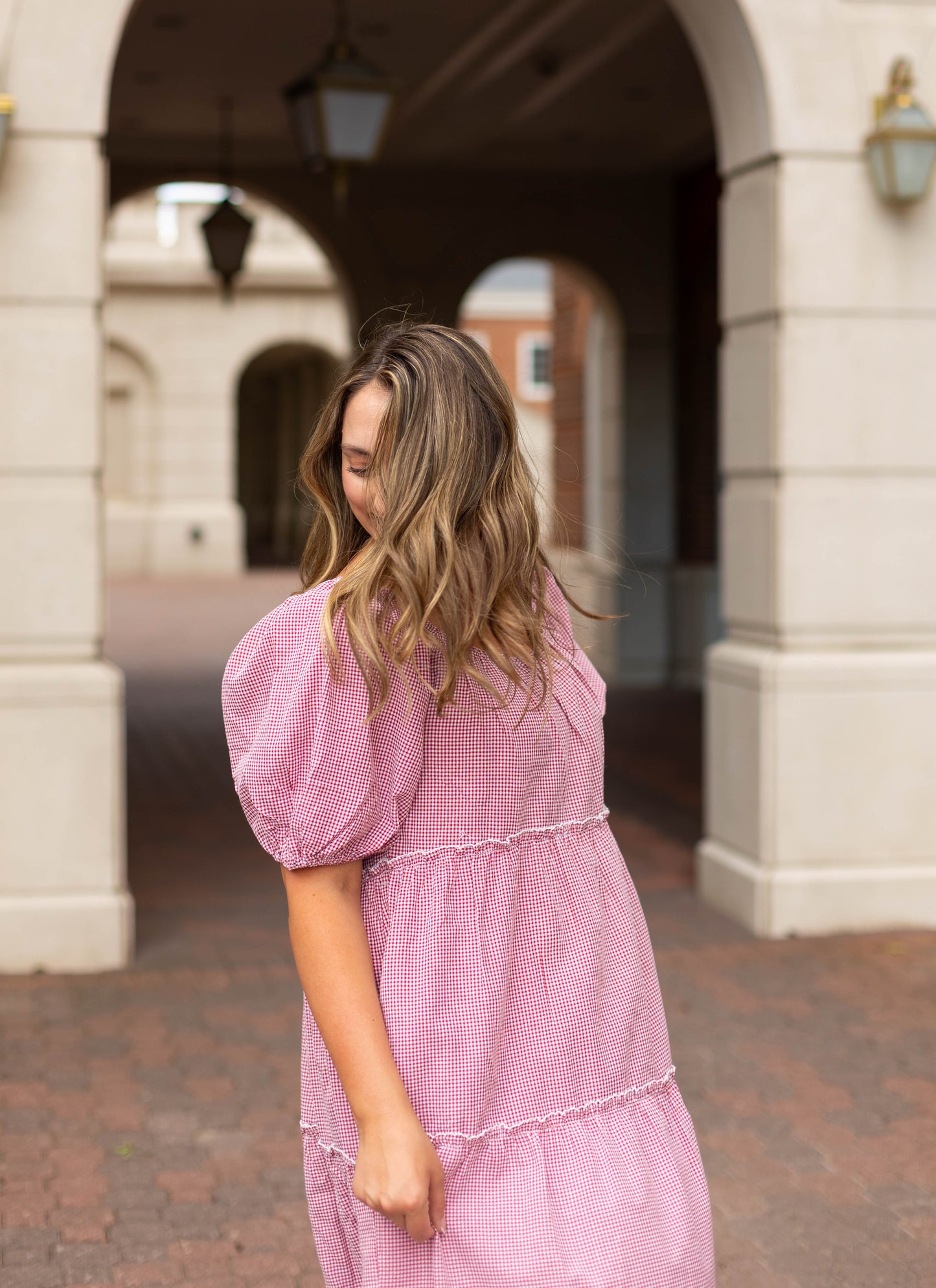 A woman wearing the Dogwood Cloth Courtlyn Tiered Gingham Midi stands outdoors, turned away from the camera beneath an archway decorated with hanging lanterns.