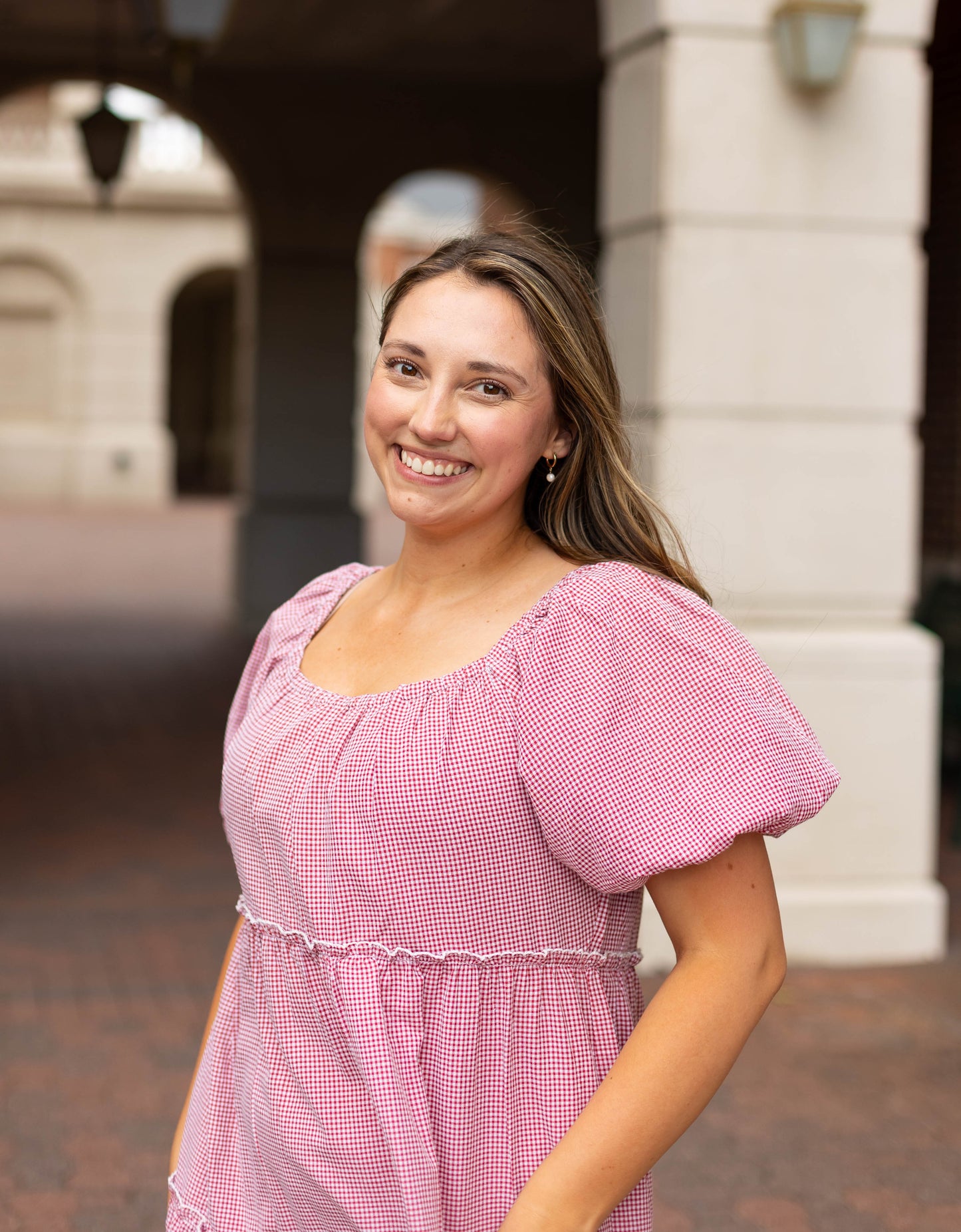 A woman with long brown hair smiles at the camera outside a building, wearing the Dogwood Cloth Courtlyn Tiered Gingham Midi dress in pink and white.