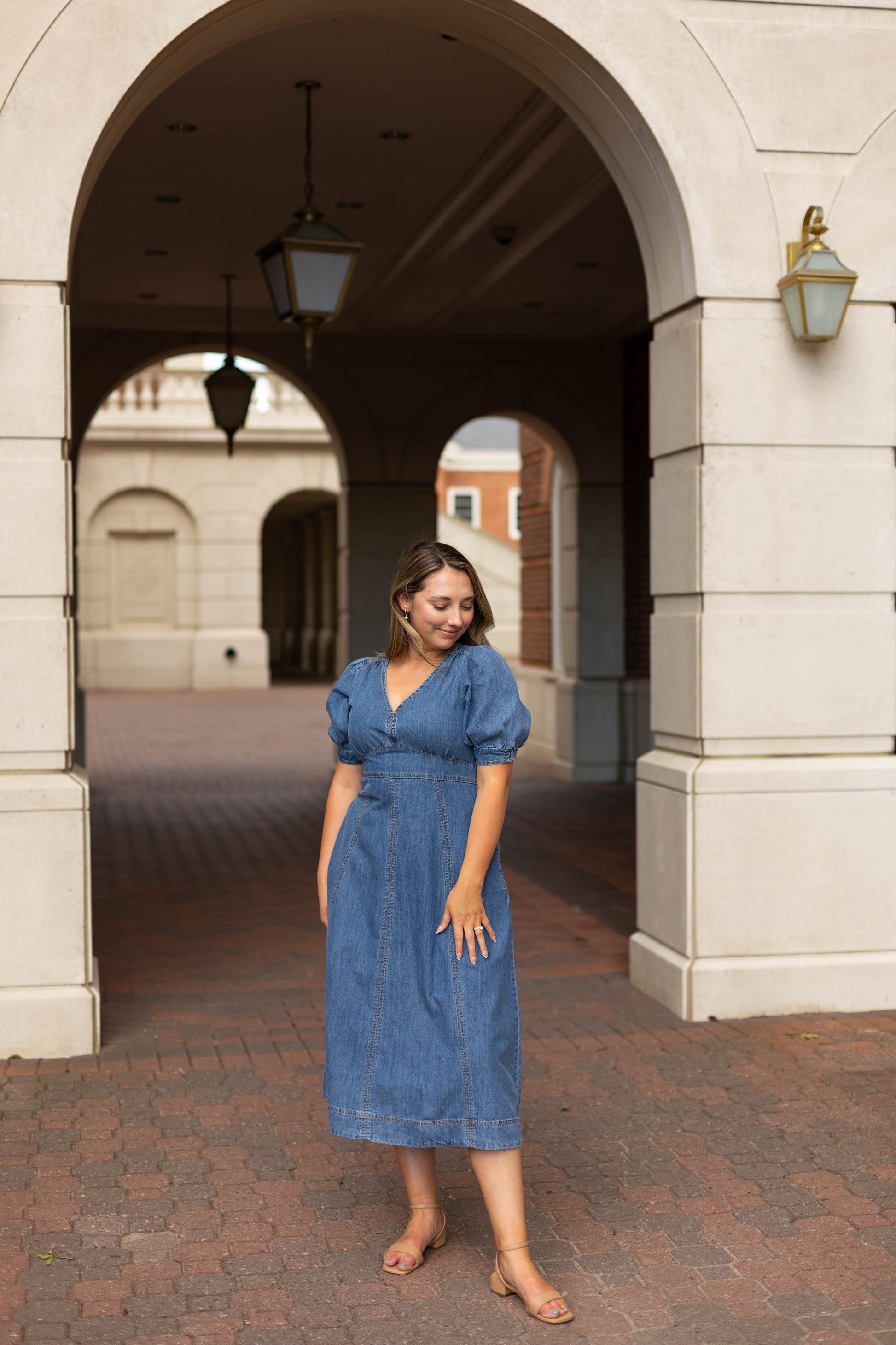 Wearing the Dogwood Cloth Kellie Denim Midi and sandals, a woman stands on a brick walkway beneath an arched stone structure with lamps.