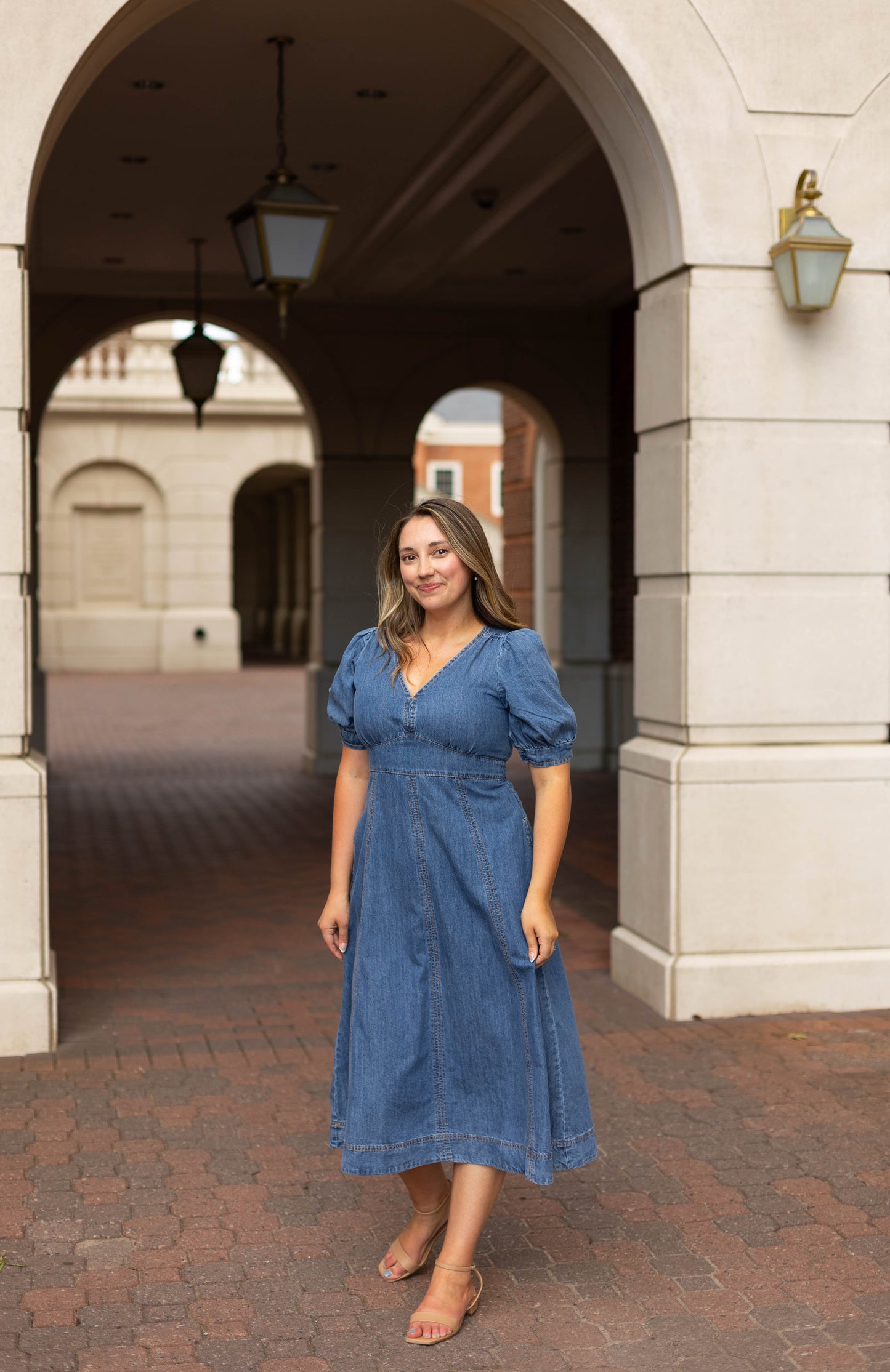 A woman wearing the Dogwood Cloth Kellie Denim Midi and sandals stands on a brick walkway under an archway of a light-colored building.