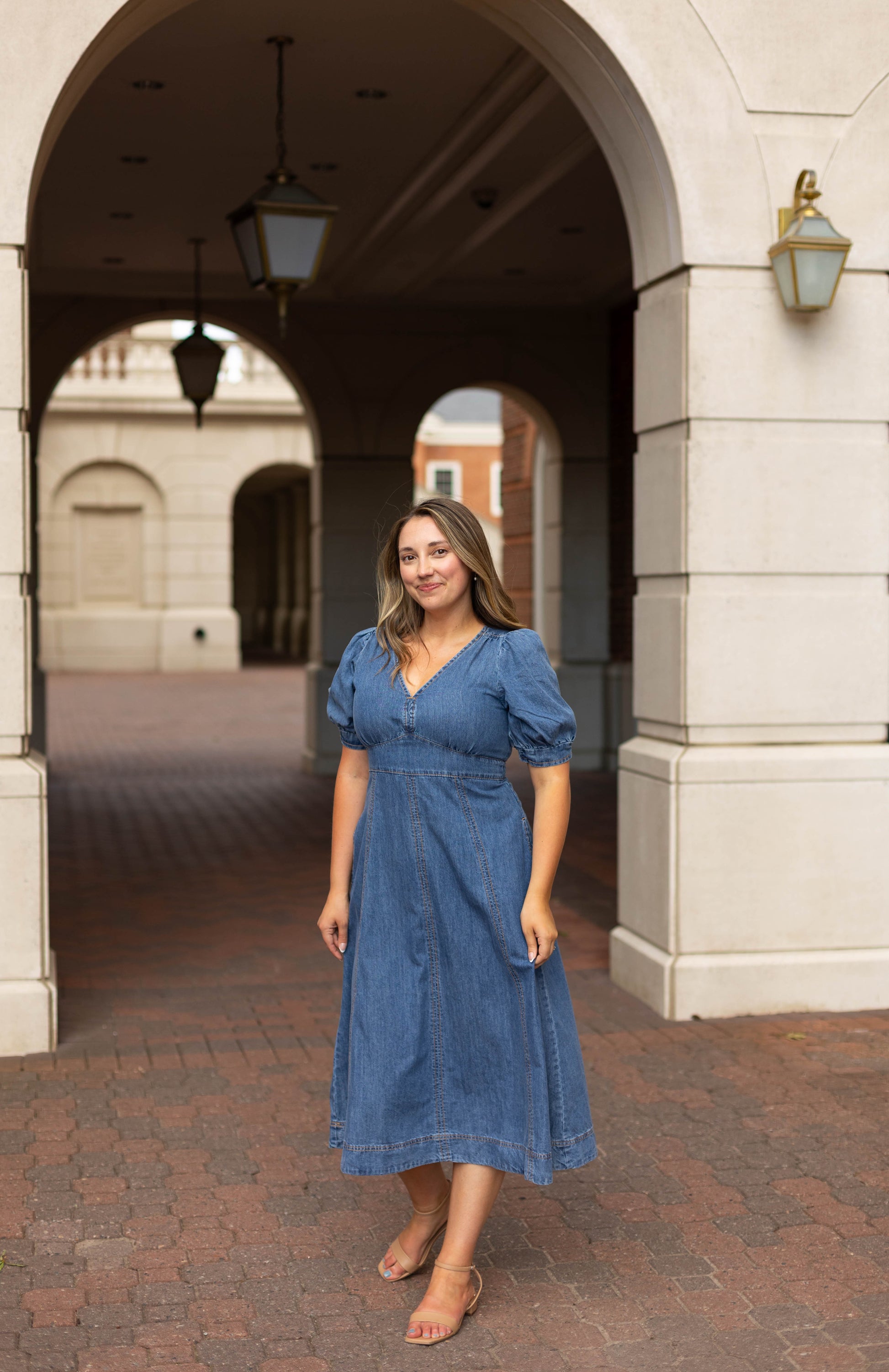 A woman wearing the Dogwood Cloth Kellie Denim Midi and sandals stands on a brick walkway under an archway of a light-colored building.