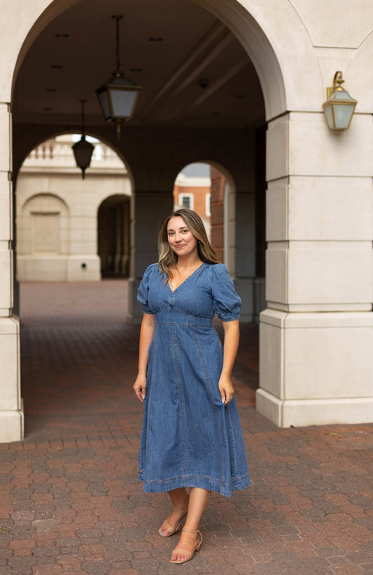 A woman wearing the Dogwood Cloth Kellie Denim Midi and sandals stands on a brick walkway under an archway of a light-colored building.