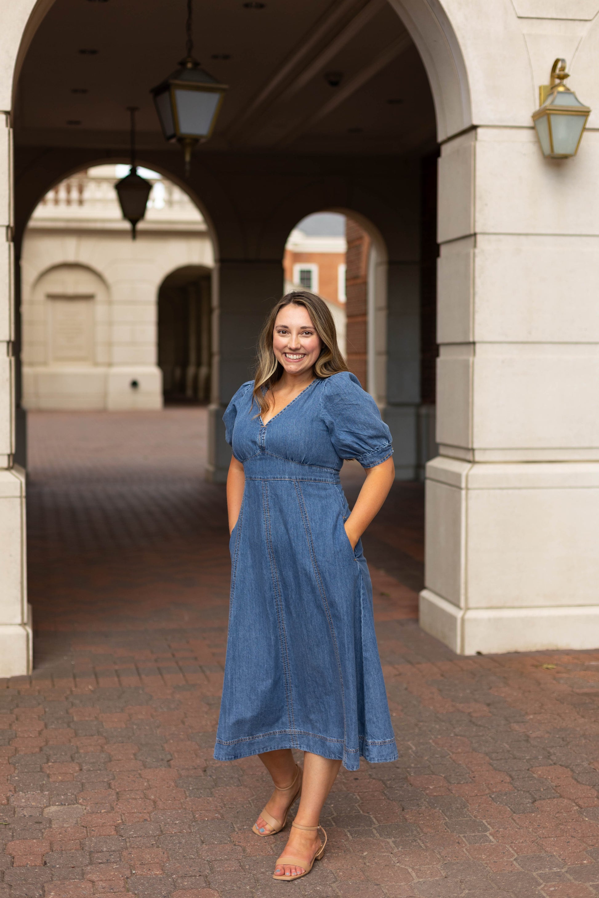 A woman smiles at the camera under an archway, hands in her pockets, wearing the Kellie Denim Midi by Dogwood Cloth.