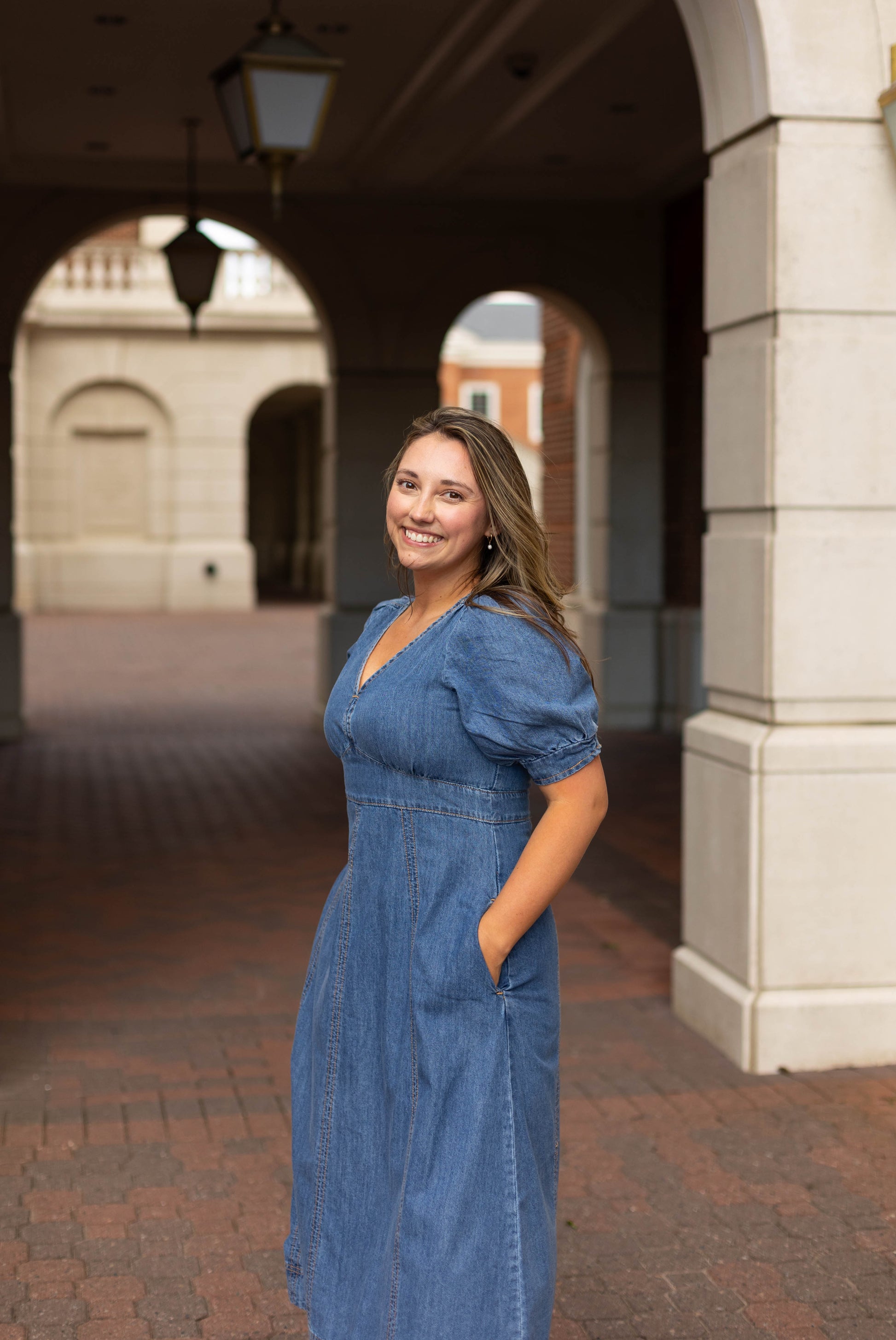 A woman wearing the Kellie Denim Midi by Dogwood Cloth stands outdoors under an archway, smiling with her hands in her pockets.