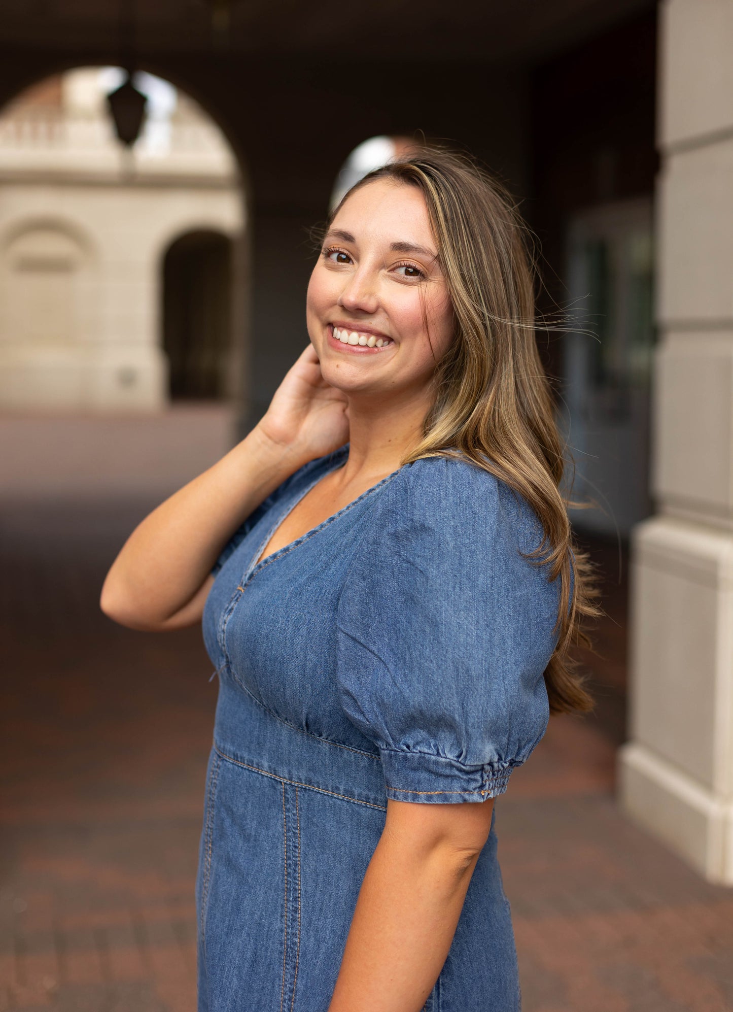 A woman with long hair smiles at the camera outdoors, wearing the Kellie Denim Midi by Dogwood Cloth and touching her neck.