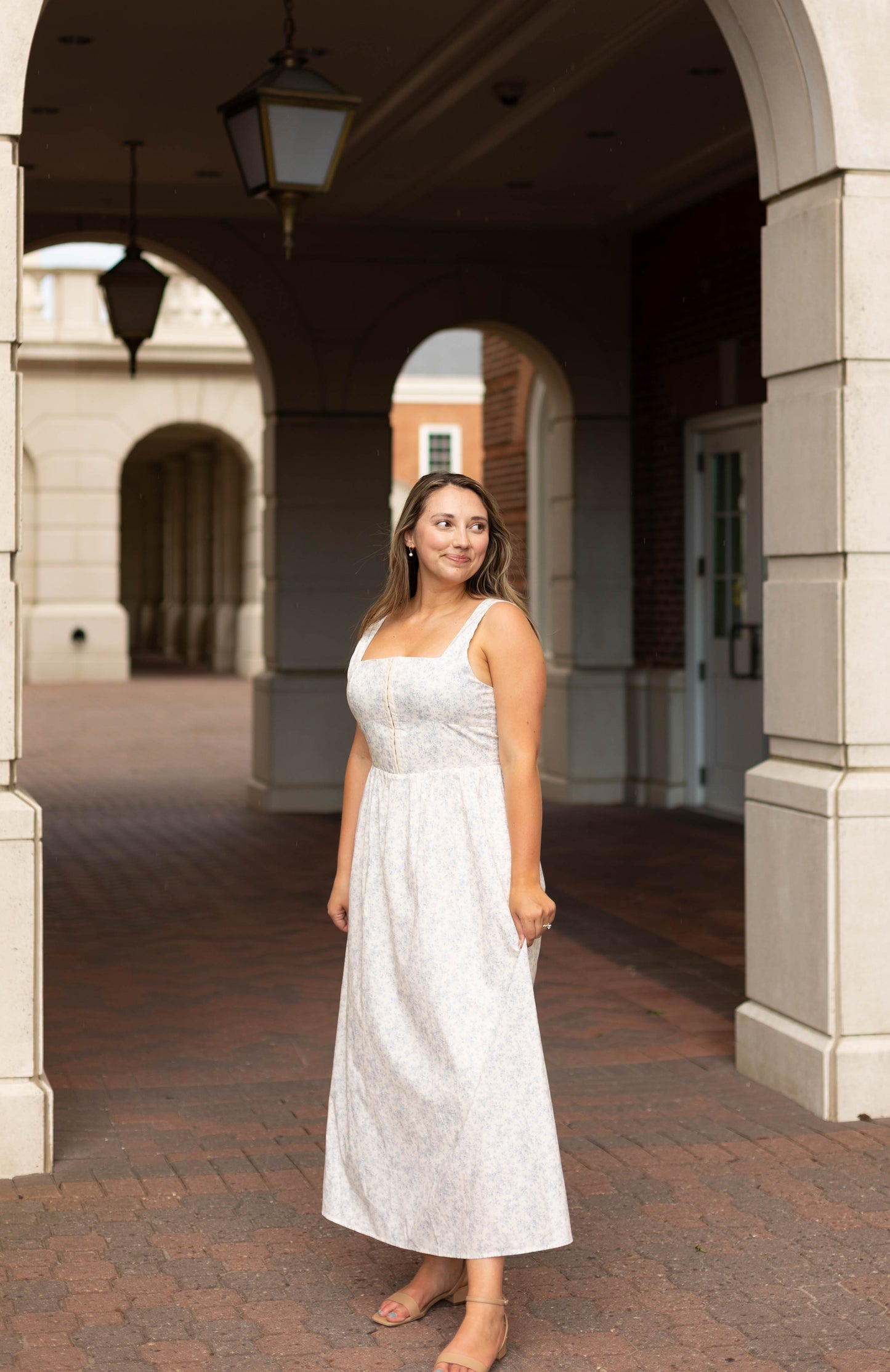 A woman in the Aurora Corset Midi by Dogwood Cloth stands under an archway in a brick courtyard, looking to her left.