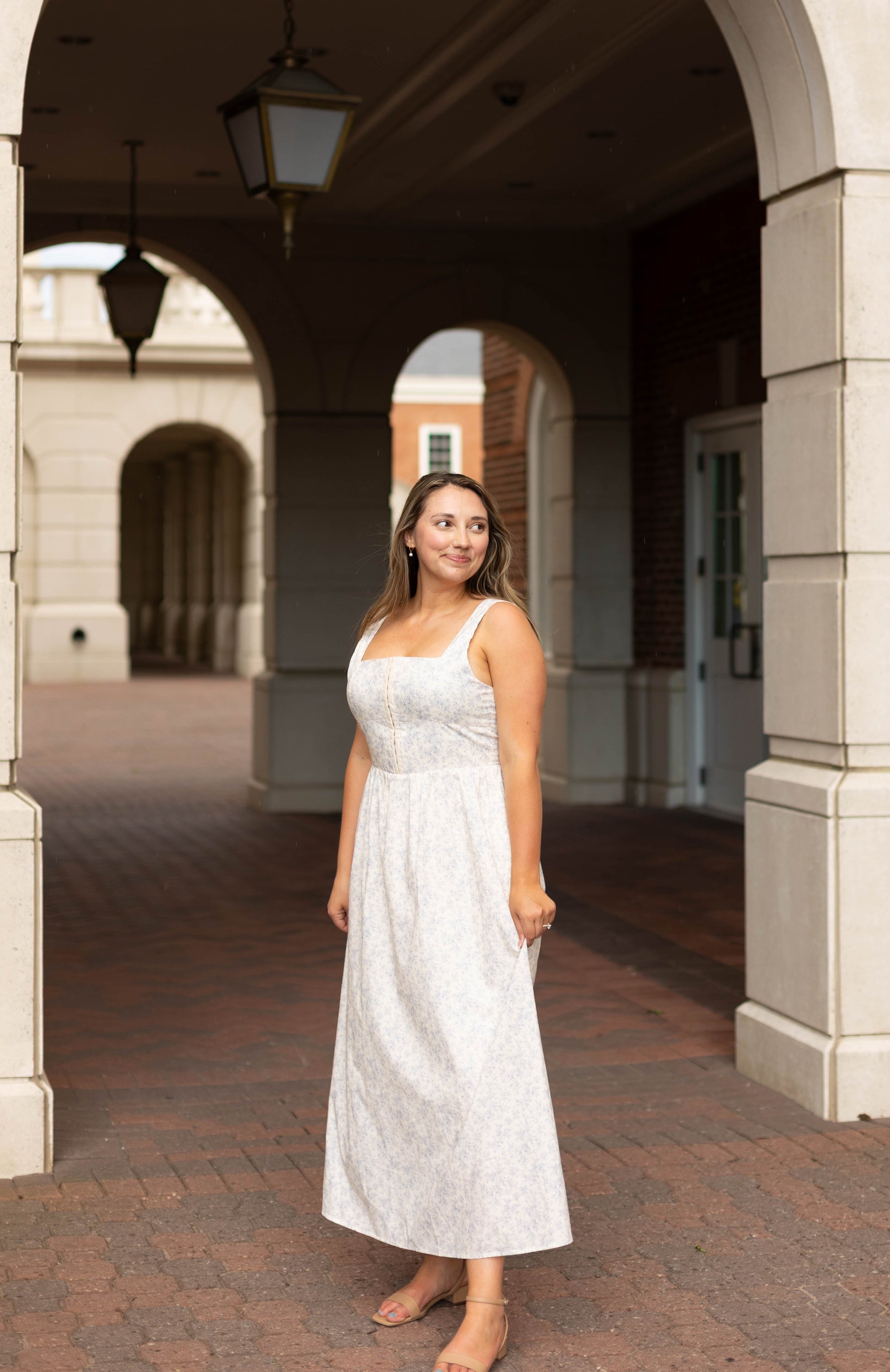 A woman in the Aurora Corset Midi by Dogwood Cloth stands under an archway in a brick courtyard, looking to her left.