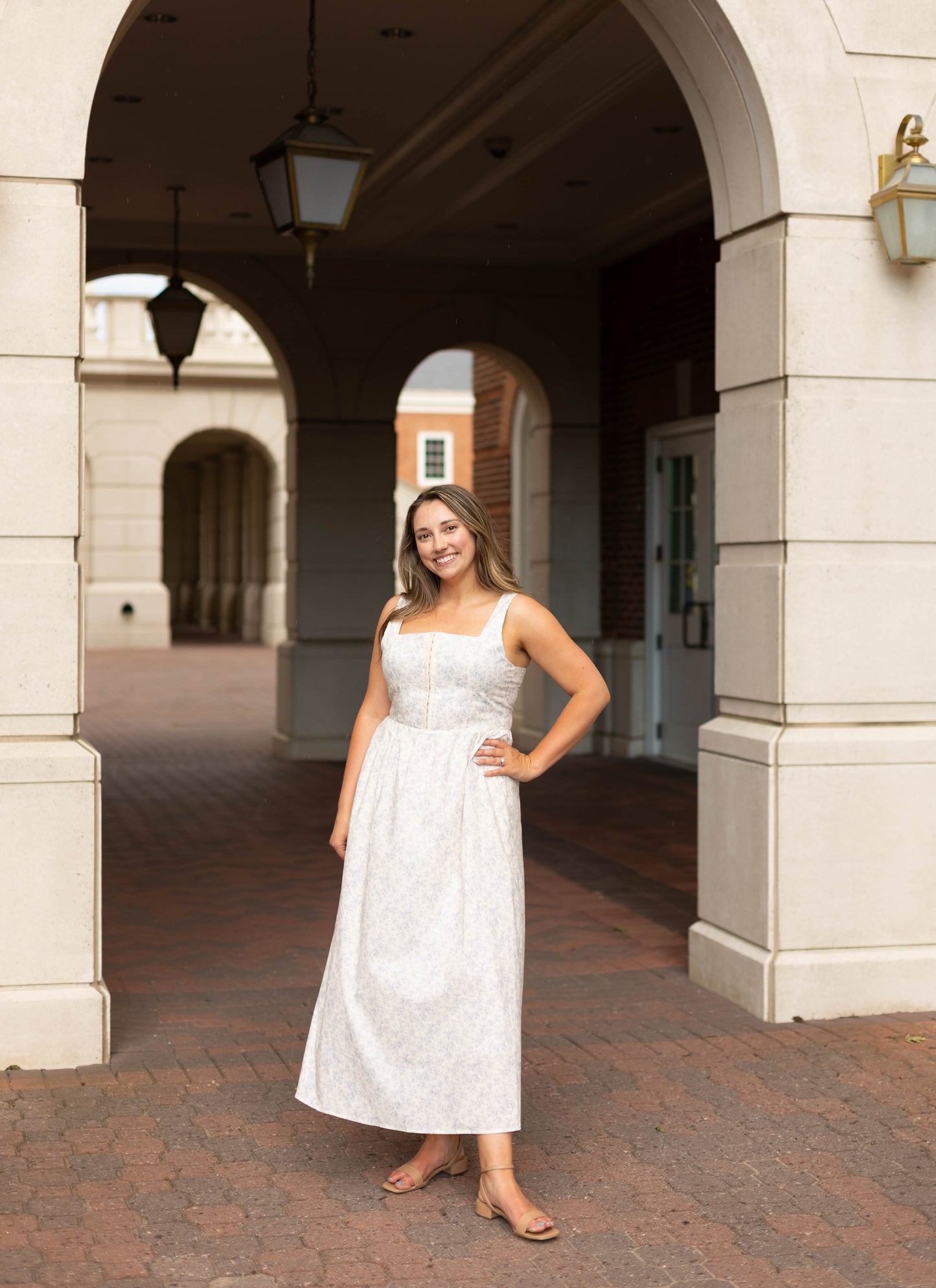 A woman smiles under a stone archway on a brick path, wearing the Aurora Corset Midi by Dogwood Cloth—a blue floral dress with a corset bodice—one hand on her hip.