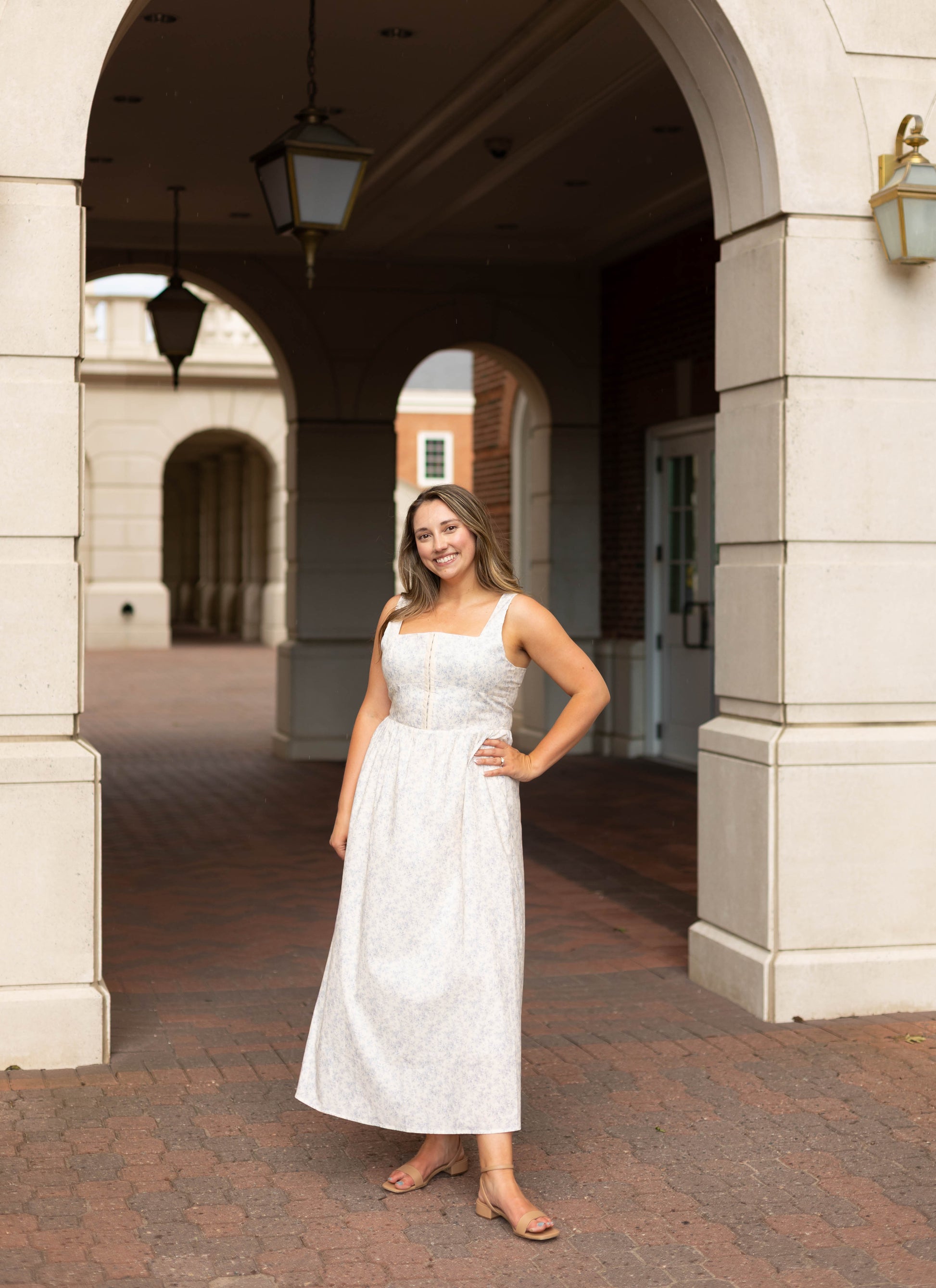 A woman smiles under a stone archway on a brick path, wearing the Aurora Corset Midi by Dogwood Cloth—a blue floral dress with a corset bodice—one hand on her hip.