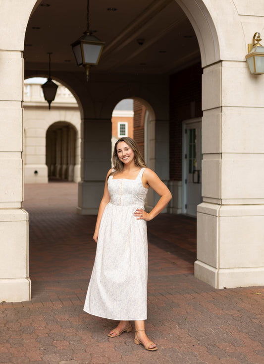 A woman smiles under a stone archway on a brick path, wearing the Aurora Corset Midi by Dogwood Cloth—a blue floral dress with a corset bodice—one hand on her hip.