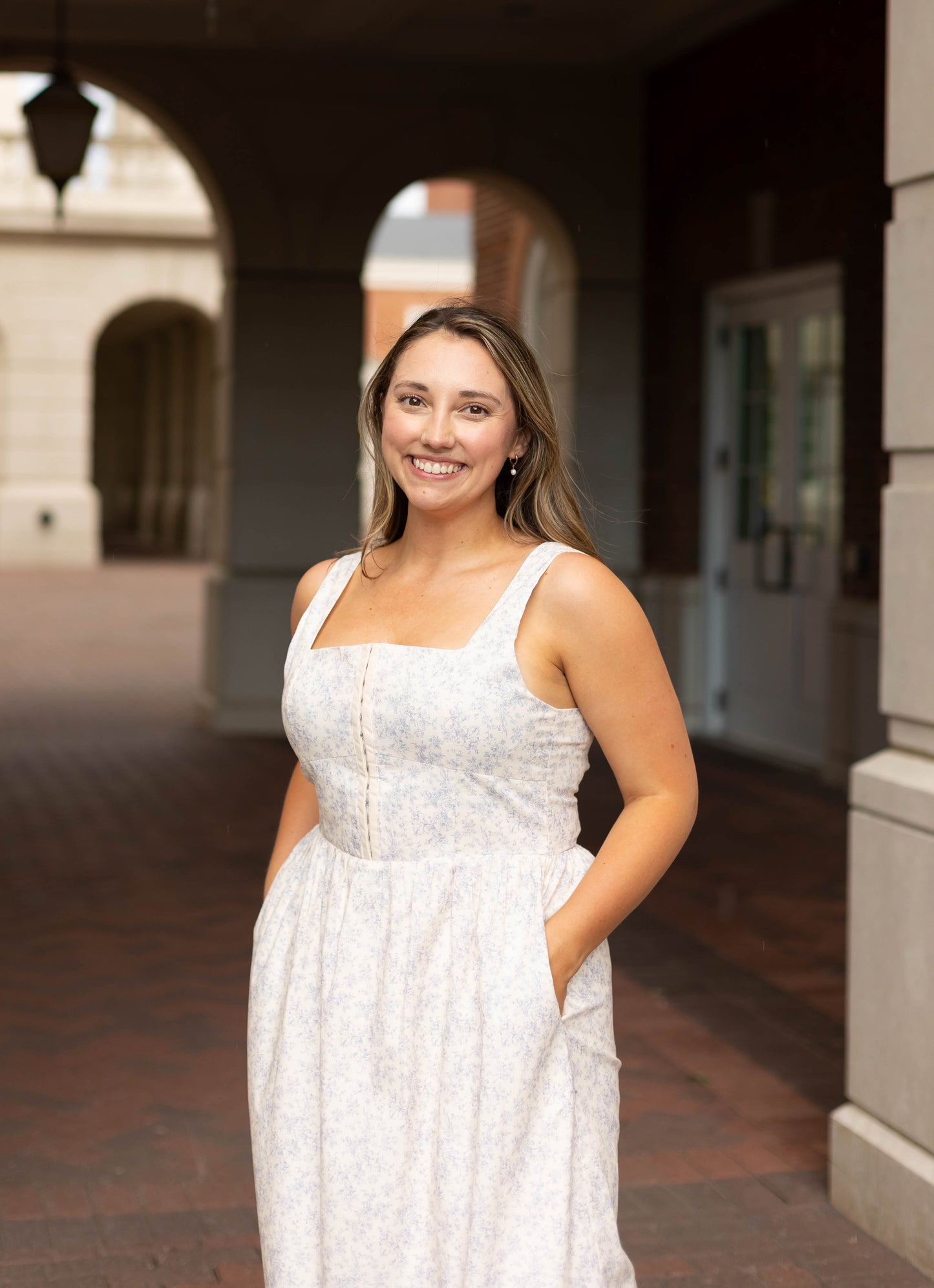 A woman wearing the Aurora Corset Midi by Dogwood Cloth stands outside under an archway, smiling at the camera with her hands in her pockets.