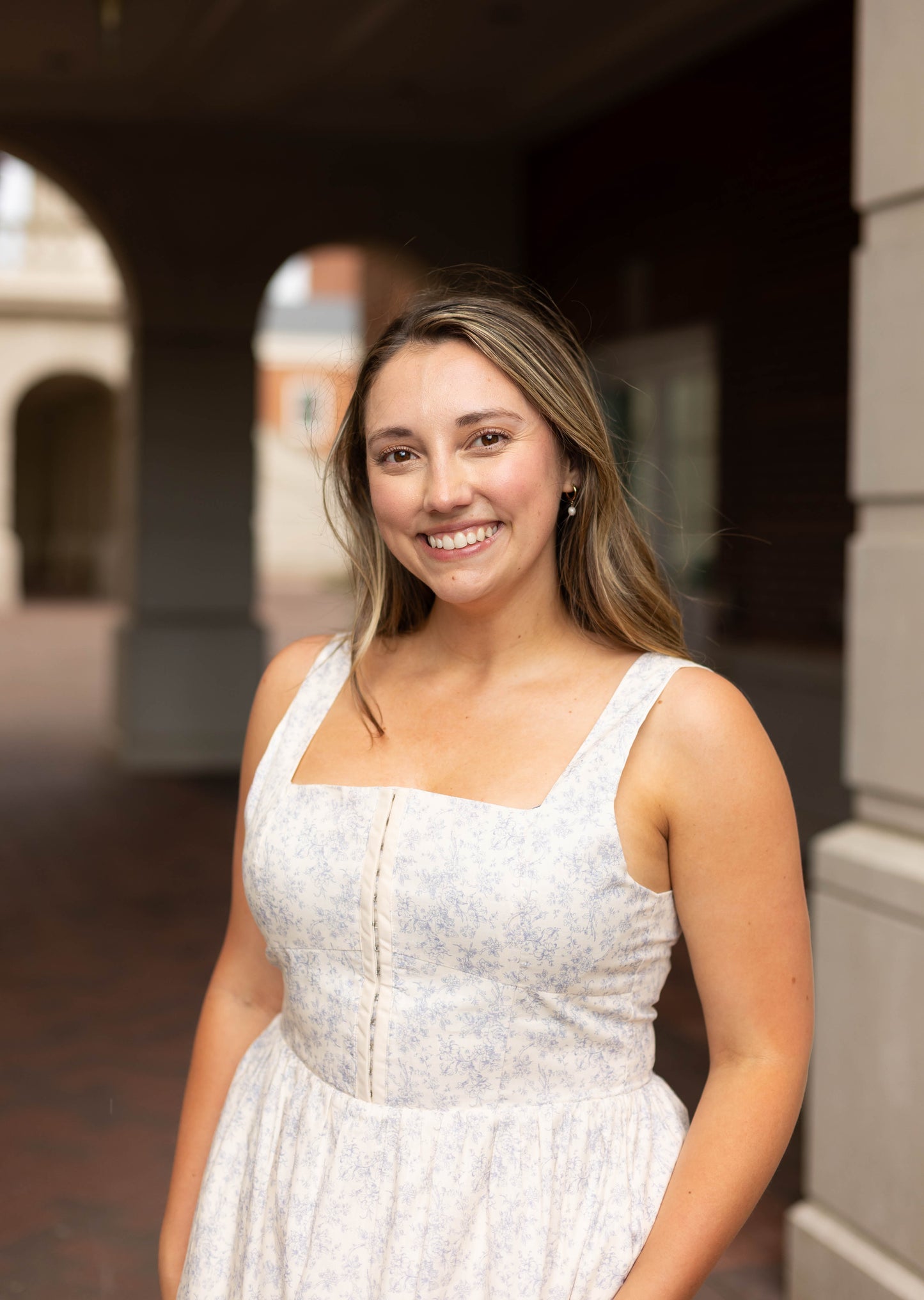 A woman with long hair smiles at the camera outdoors in a covered walkway, wearing the Aurora Corset Midi by Dogwood Cloth.