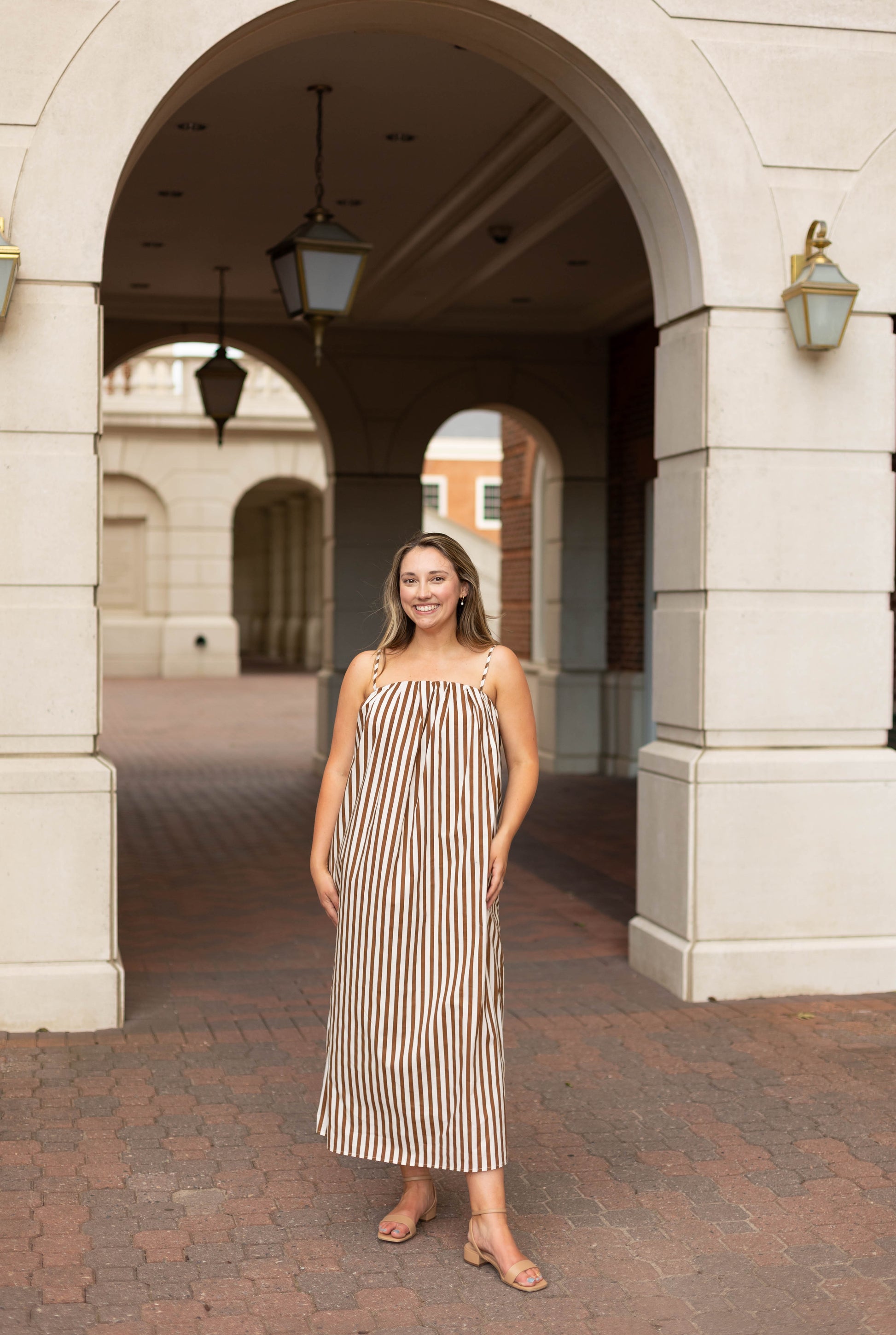 A woman in the Dogwood Cloth Jeanine Striped Maxi, a brown and white dress with adjustable straps, smiles while standing under a stone archway on a brick pathway.