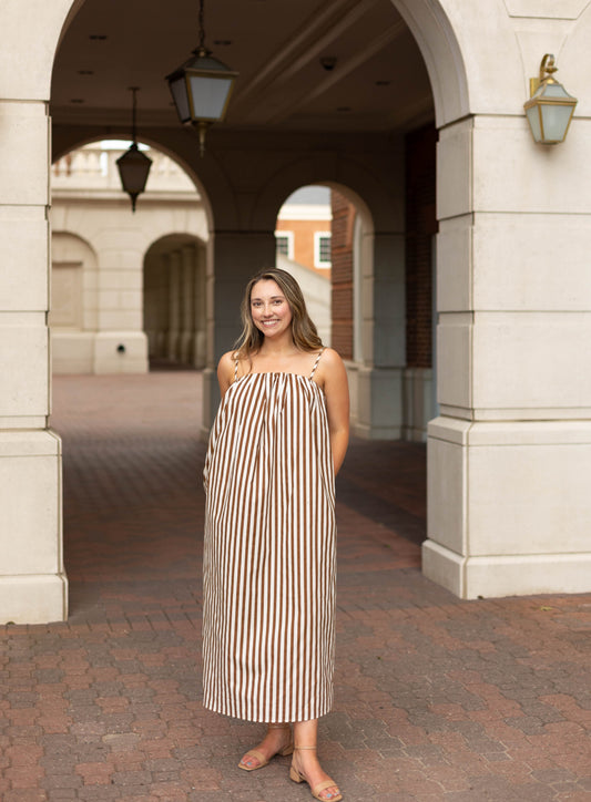 A woman stands outside on a brick walkway wearing the Dogwood Cloth Jeanine Striped Midi in mocha and cream, with sandals and hands in pockets. Arched architectural features appear in the background.