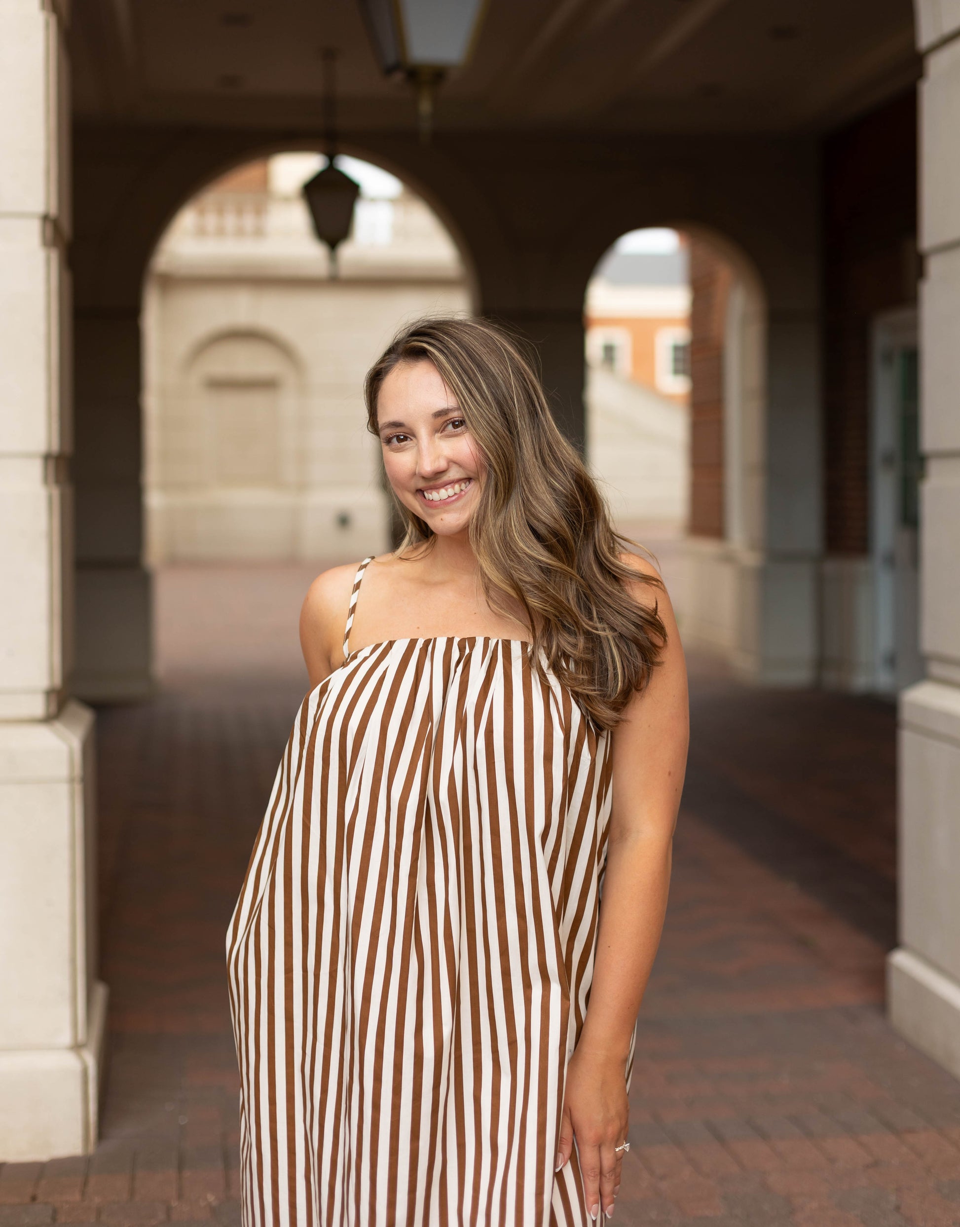 A woman smiles under an archway in an outdoor corridor, wearing the Dogwood Cloth Jeanine Striped Maxi—a brown and white striped dress with adjustable straps.
