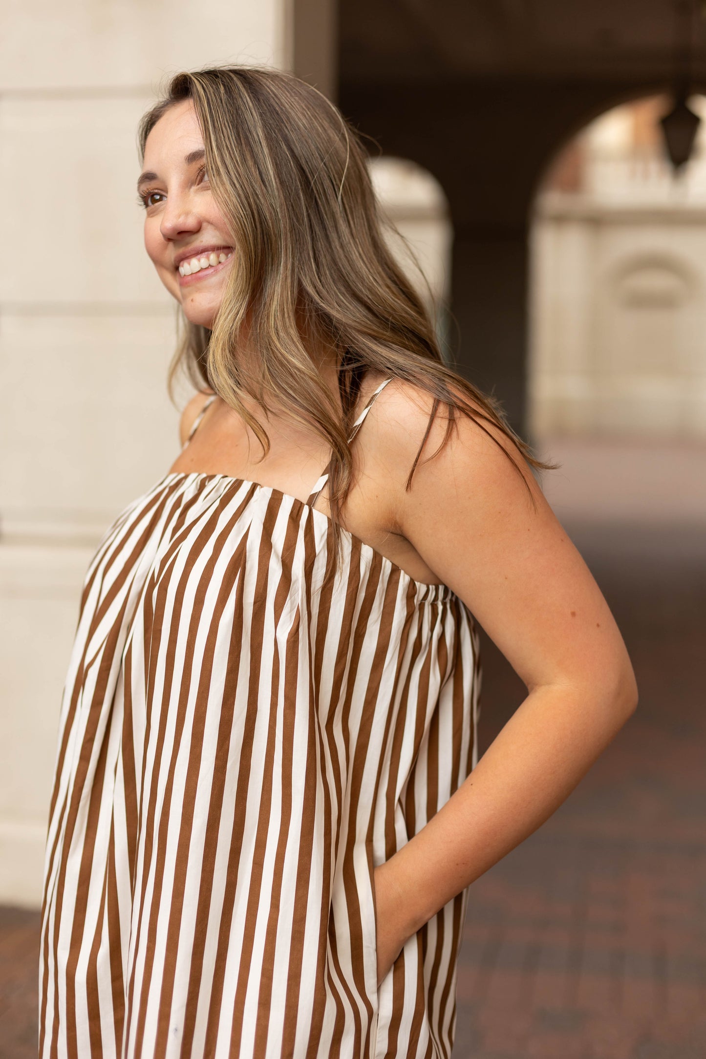 A woman stands outdoors, smiling, with her hand in her pocket, wearing the Dogwood Cloth Jeanine Striped Maxi—a brown and white striped dress with adjustable straps.