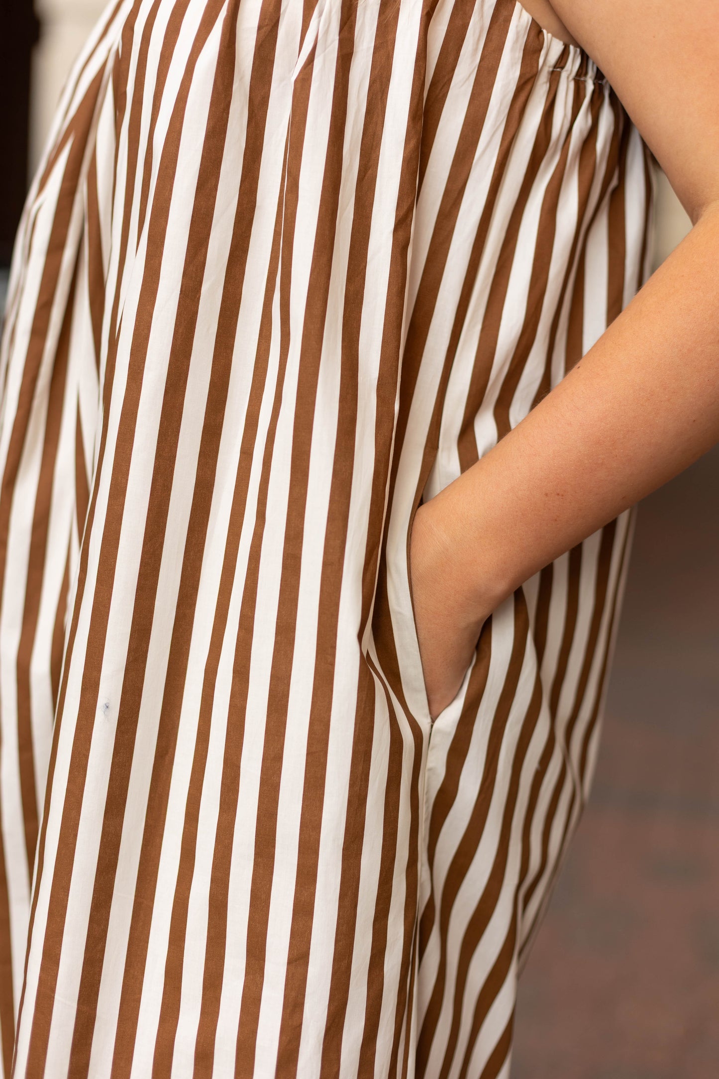 Person wearing the Dogwood Cloth Jeanine Striped Maxi, a brown and white vertically striped dress with adjustable straps, right hand tucked into a side pocket.