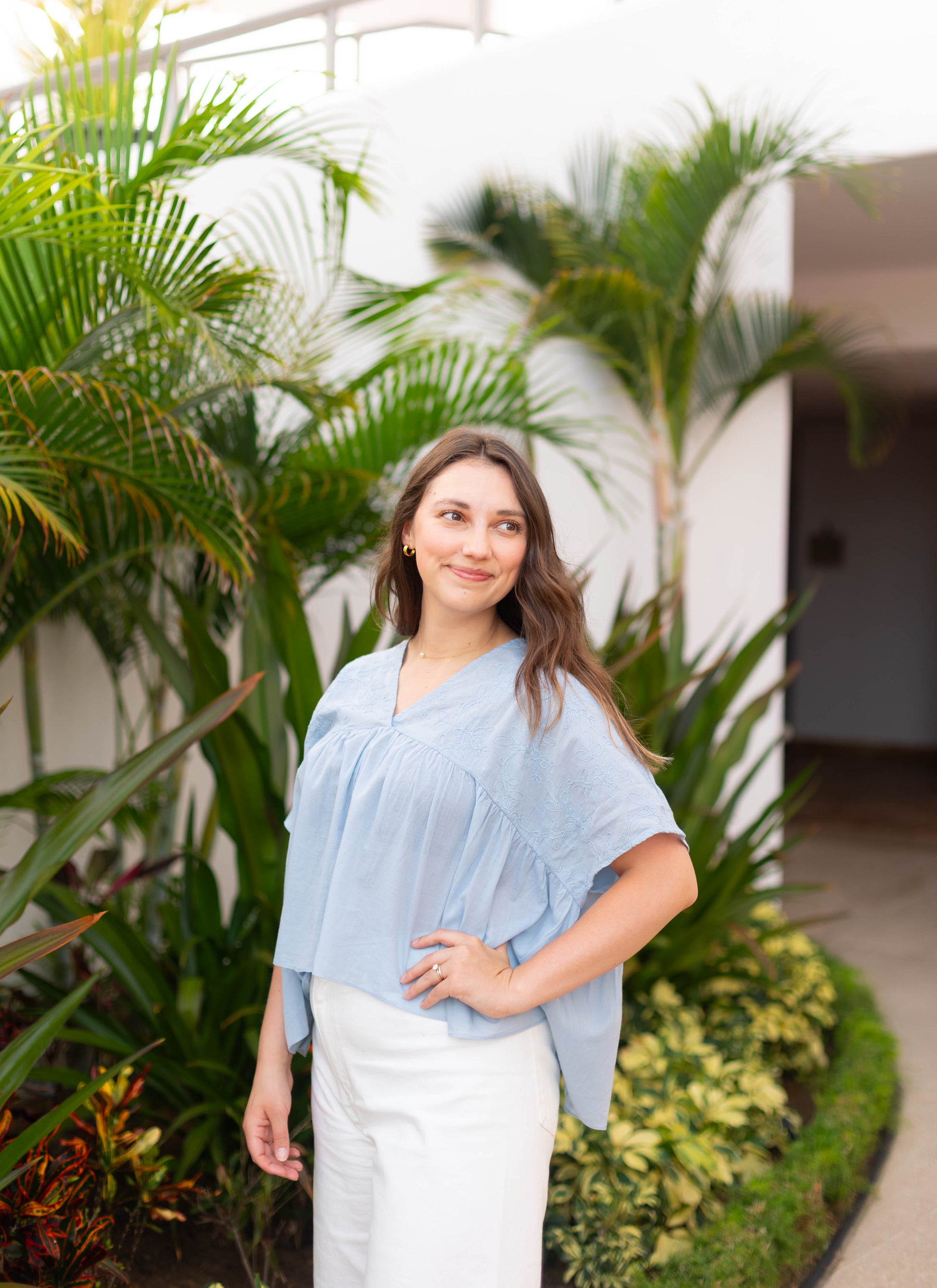 A woman with long brown hair stands outdoors near green plants, smiling and looking to the side. She wears the Francesca Top by Dogwood Cloth, a light blue blouse with embroidered detailing, paired with white pants.