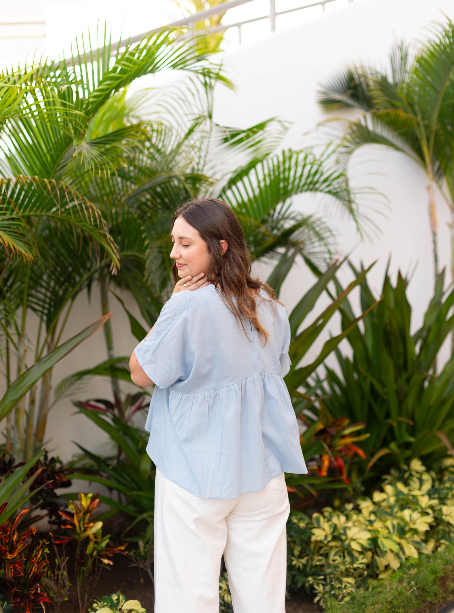 A woman with long hair stands outdoors before green plants, wearing Dogwood Cloth’s Francesca Top in a relaxed fit light blue style paired with white pants, as she looks over her shoulder.