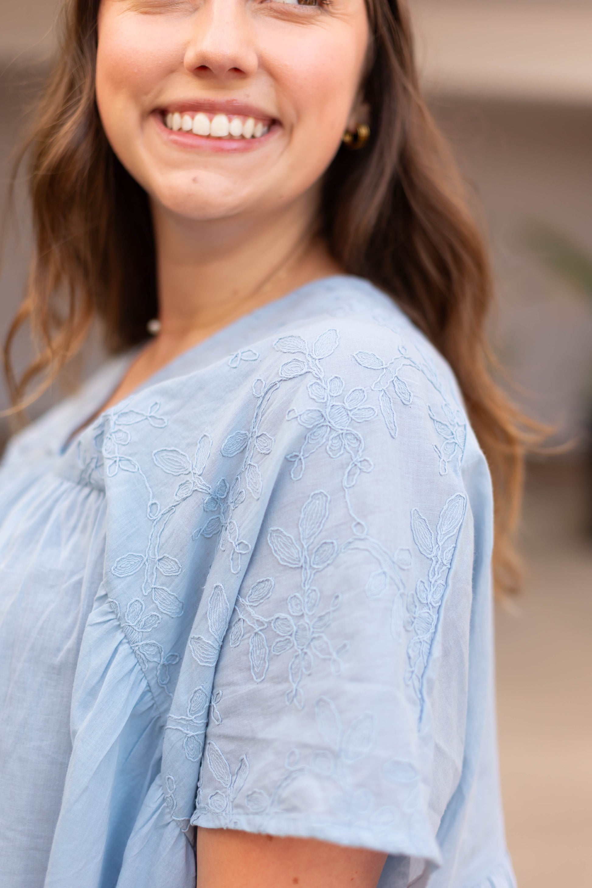 A woman smiles, shown from the shoulders up with her face partially out of frame, wearing Dogwood Cloth’s The Francesca Top in a relaxed fit and light blue with embroidered detailing.