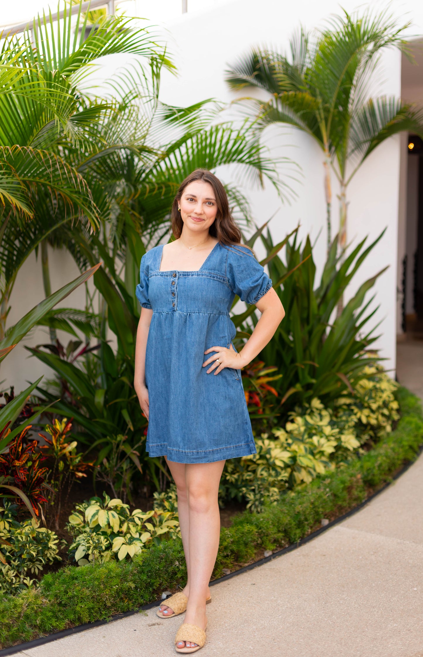 A woman models The Taylor Denim Dress by Dogwood Cloth, featuring puff sleeves, as she poses with one hand on her hip in front of lush tropical greenery.