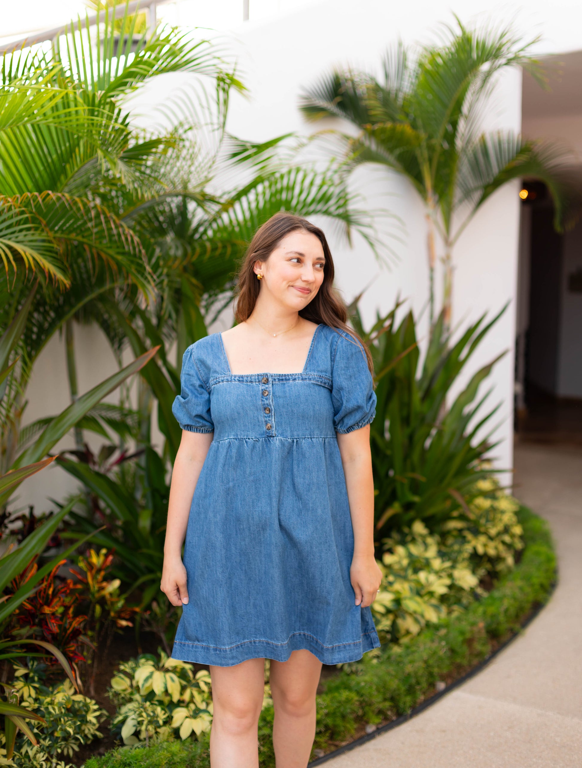 A woman wearing the Dogwood Cloth Taylor Denim Dress with puff sleeves stands on a garden path, surrounded by green plants and palm trees, gazing to the side with a slight smile.