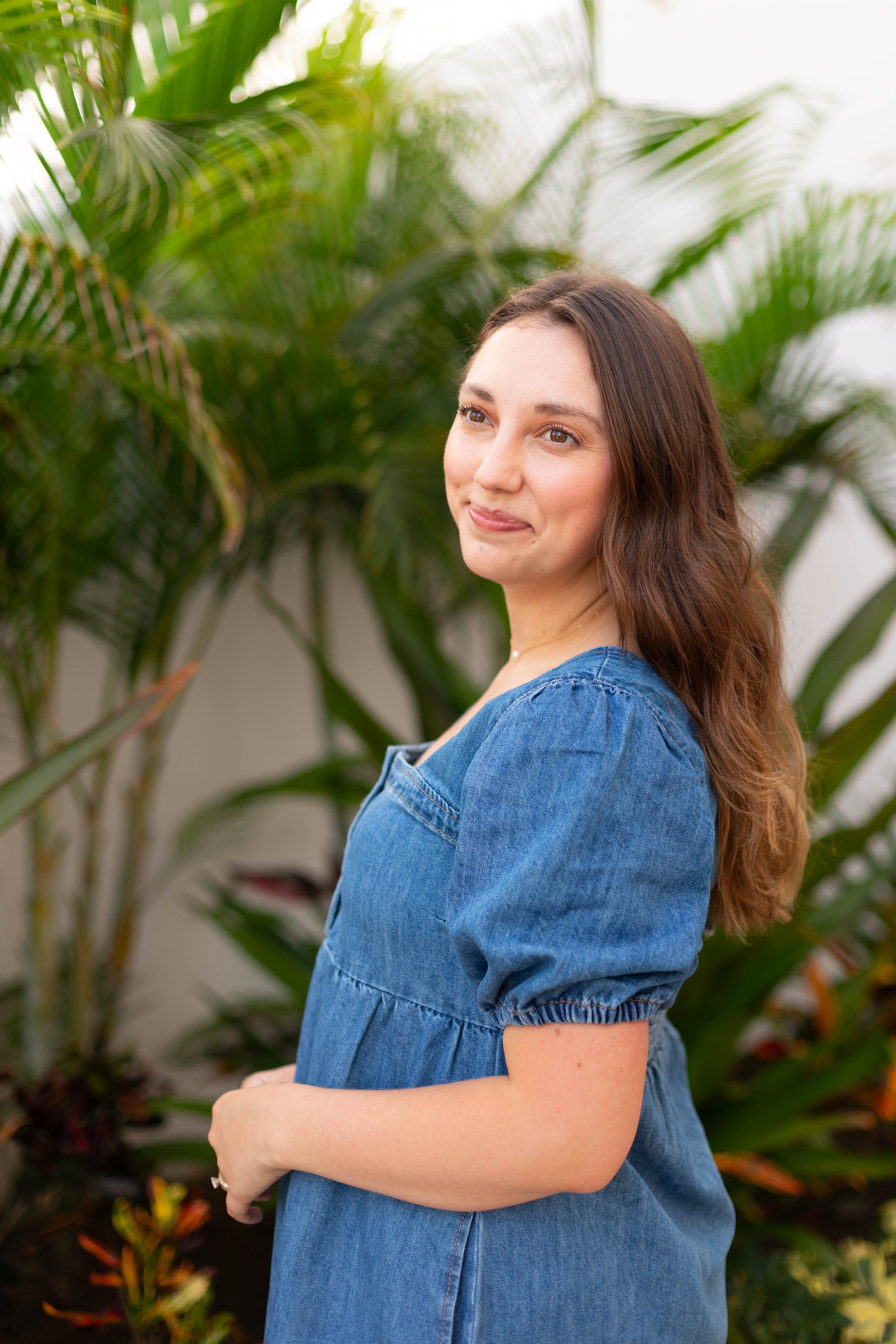 A woman with long brown hair wears the Taylor Denim Dress by Dogwood Cloth, featuring a square neckline, while standing outdoors among green leafy plants and softly smiling over her shoulder.